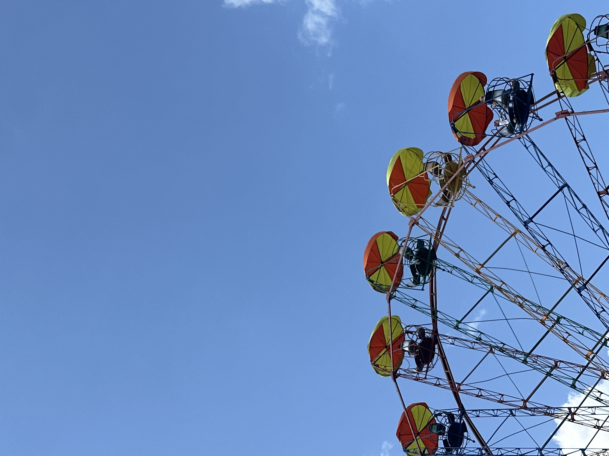 The image shows a Ferris wheel against a clear blue sky with a few scattered clouds. The Ferris wheel is partially visible on the right side of the frame. The gondolas have a distinctive design with a combination of red and yellow panels. The Ferris wheel's structure is made of metal beams forming a network of triangles. The overall scene suggests an amusement park. There is no indication of people or other subjects in the scene.