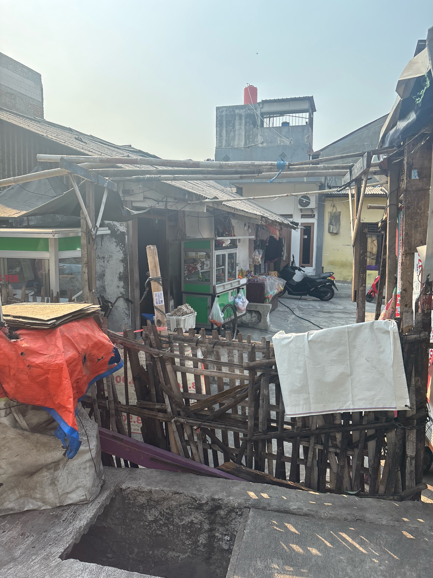 The image depicts a street scene in a densely populated urban area, likely a slum or informal settlement, in North Jakarta, Indonesia. The architecture is a mix of makeshift structures with corrugated metal roofs and more permanent buildings. There is evidence of commercial activity with small roadside stalls or shops. In the foreground, a wooden fence made of uneven planks encloses an area with various items, including large sacks, a stack of flat boards covered by an orange tarp, and a white fabric sheet hanging on the fence. A partially visible concrete structure with a hole in it is also in the foreground. Behind the fence, a street or open area is visible, with a motorcycle parked and a small red scooter further in the background. Several buildings are visible, including one with a red water tank on the roof. The overall impression is one of a busy, somewhat cluttered urban environment.