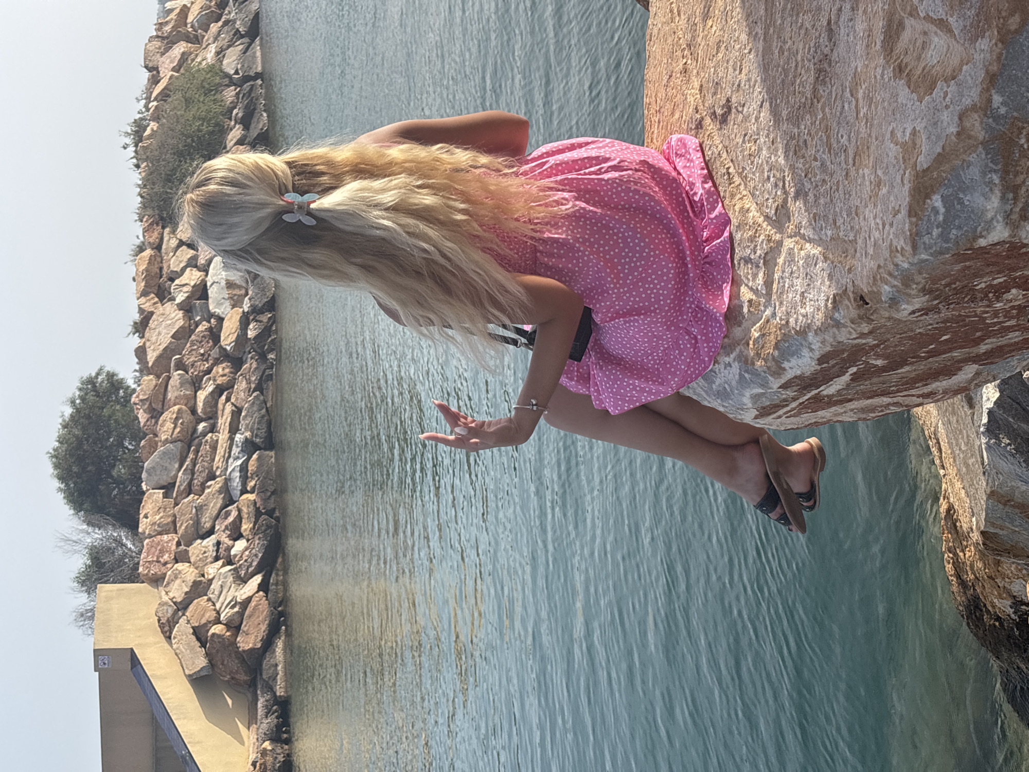 A woman with blonde hair tied back with a light blue and red butterfly clip sits on a large rock overlooking a body of water. She is wearing a short pink dress with white polka dots and black sandals. Her right hand is raised with her fingers extended. The water is calm and reflects the light from the sky. Behind her, a rocky embankment is visible, with some trees and a light-colored structure in the background. The overall scene suggests a sunny day at a coastal location.
