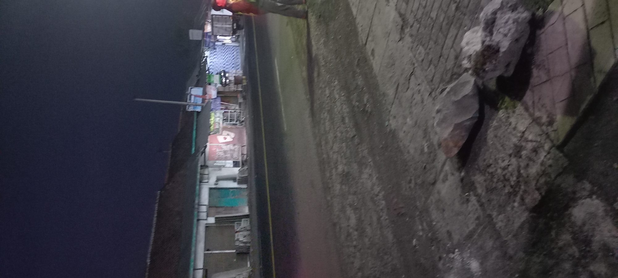 The image depicts a street scene at night in Banjarsari, Indonesia. In the foreground, large rocks are visible on a paved surface. A road runs parallel to a row of buildings on the left side of the image, with a yellow line marking the edge of the road. The buildings appear to be shops or businesses, with shutters down on some of them. Signs are visible on the storefronts, though they are illegible due to the low light and image quality. A person wearing a high-visibility vest and a red head covering stands on the right side of the road, facing away from the camera. The sky is dark, indicating nighttime. The overall scene suggests a quiet street with some commercial activity or infrastructure visible.