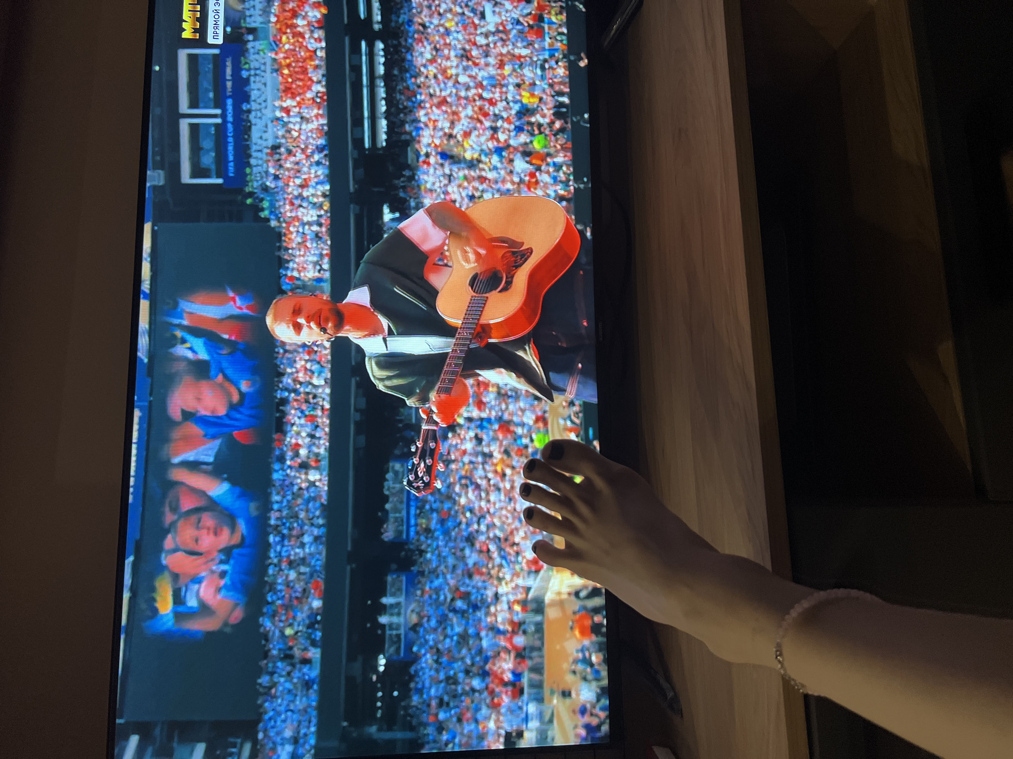 A full-frame, eye-level shot shows a television screen displaying a performance with a person playing an acoustic guitar. The screen also features a large crowd of spectators in the background. In the foreground, a left foot with dark nail polish and a bracelet is placed on a wooden surface. The television is placed on a dark wooden media console. The scene appears to be indoors, likely a living room, with no specific identifying markers to pinpoint a location in Moscow, Russia.