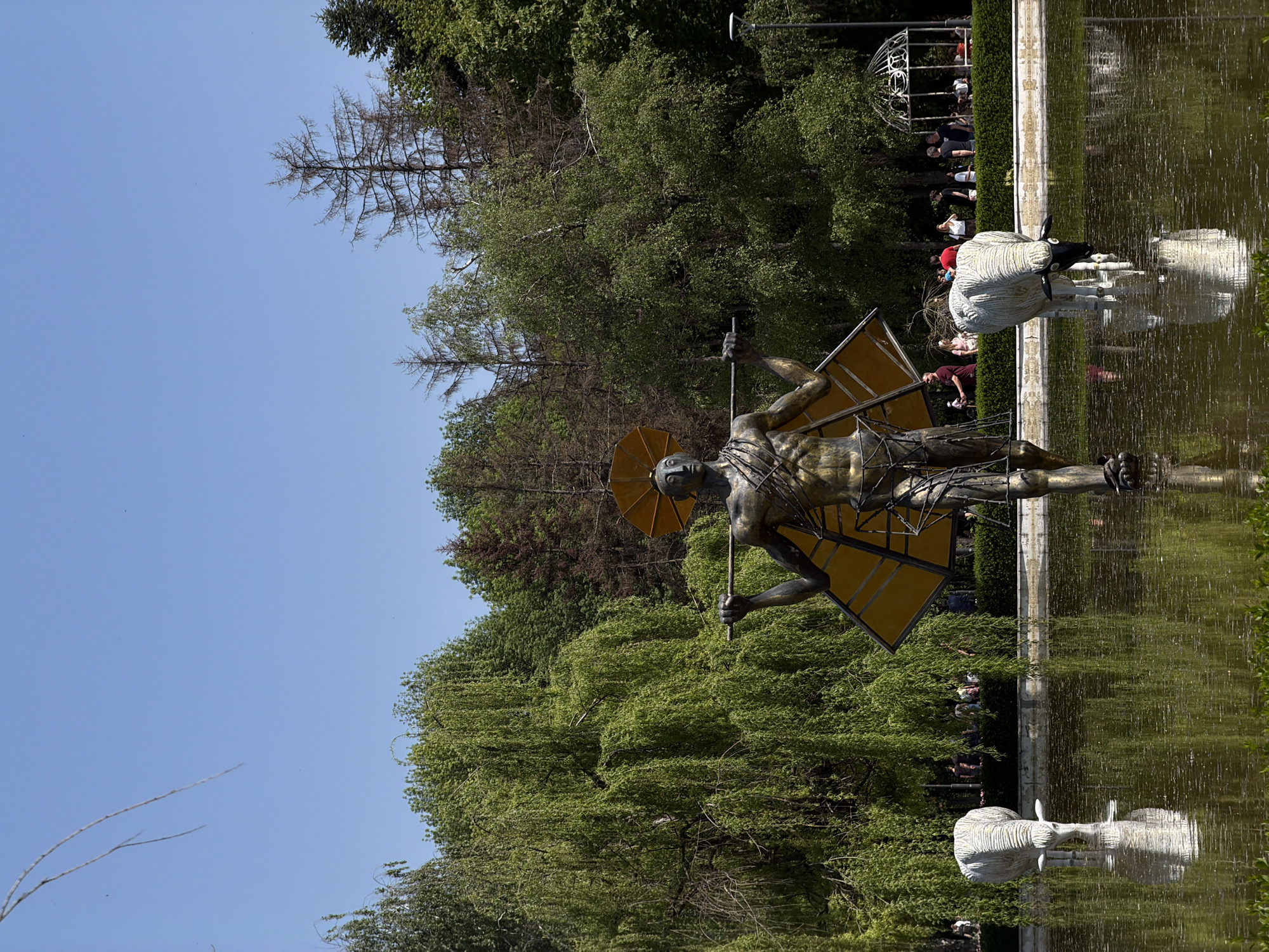 The image depicts a vertical, outdoor scene. A bronze statue of a male figure, seemingly Icarus, stands in a shallow pool of water. The statue has a yellow rectangular wing structure. The reflection of the statue is visible in the water. Beside the statue is a white ram statue, also reflected in the water. Lush green trees form the backdrop. In the background, people are present near a white gazebo structure. The sky is clear blue. The location appears to be a park in Sofia, Bulgaria.