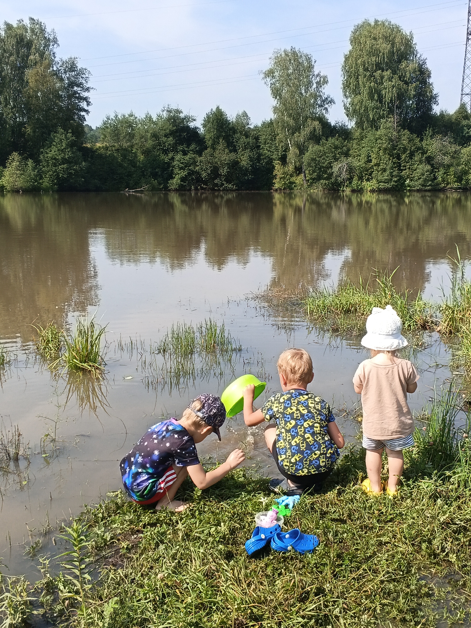 Three young children are playing by the edge of a muddy body of water. The child on the left is wearing a dark t-shirt with a galaxy print and a cap, and is partially submerged in the water, holding a green bucket. In the center, another child in a patterned t-shirt is also crouched by the water's edge, holding a green shovel. The child on the right, wearing a tan shirt and striped shorts, stands facing the water, wearing a white bucket hat and yellow rain boots. A pair of blue Crocs and some small toys are on the grassy bank. The background consists of lush green trees and a blue sky with a few power lines visible.