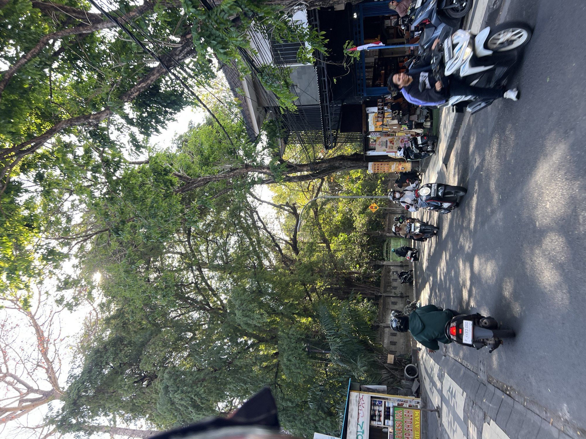 In Kuta, Indonesia, a street scene unfolds with a clear view of a road lined with lush green trees and a building with multiple levels. The sun shines through the foliage, casting dappled shadows on the asphalt. Several individuals are riding motorcycles, navigating the roadway. In the foreground, a person on a white motorcycle is captured mid-motion, smiling. Behind them, another individual on a dark motorcycle, wearing a green hoodie, is also riding. Further back, a line of motorcycles continues down the street. Shops with various merchandise are visible along the side of the road, one displaying a sign that reads "BAKSO IGA MR. SHOLEH MIE AYAM." The architecture of the building suggests a commercial establishment, with balconies and numerous electrical wires strung across its facade. The overall atmosphere is one of everyday activity in a bustling urban environment.