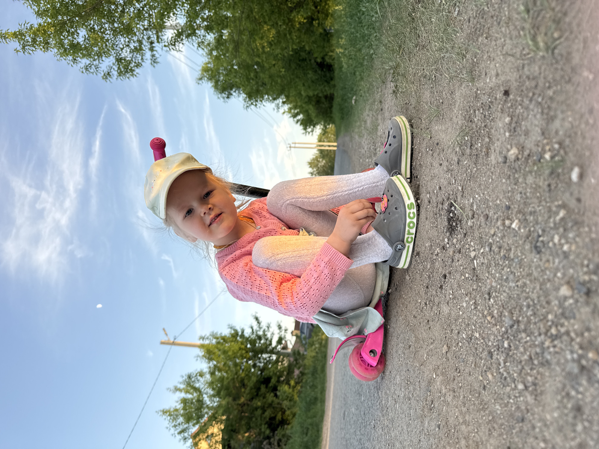 A young girl is sitting on a pink scooter on an asphalt path in Горный, Russia. She is wearing a light-colored cap with a pink object on top, a pink sweater, white leggings, and gray Crocs shoes. The path is bordered by greenery and trees. The sky is visible with some clouds and a moon.