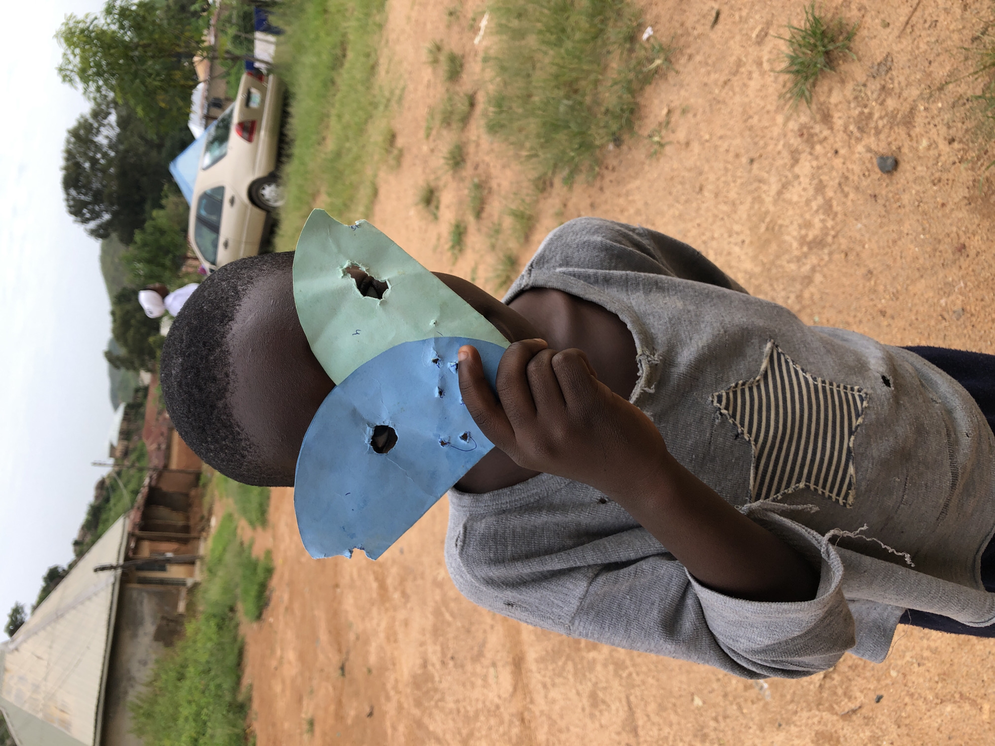 A close-up, medium shot captures a young child partially obscuring their face with two pieces of torn, colored paper, one blue and one light green, both with circular holes punched through them. The child, who appears to be of African descent, is wearing a grey, long-sleeved shirt with frayed edges and a star-shaped patch on the chest. The background suggests a rural or semi-rural setting. To the left, a simple building with a metal, gabled roof is visible, surrounded by sparse vegetation. Further back, a light-colored car is parked on a dirt road, with trees and hills in the distance under an overcast sky. This scene is consistent with the outskirts of the city of Jos, Nigeria, characterized by its reddish soil, modest structures, and surrounding natural landscape.