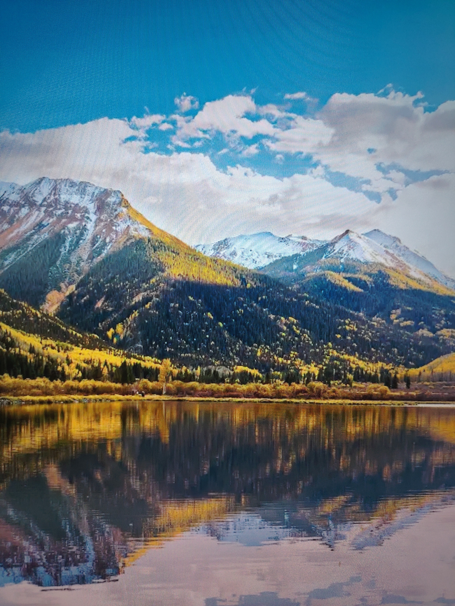 The image depicts a mountain range reflected in a body of water. The mountains are covered in a mix of green trees, yellow foliage, and snow-capped peaks. The sky is blue with scattered white clouds. The reflection in the water mirrors the mountains and sky. The location is Unknown, Unknown.