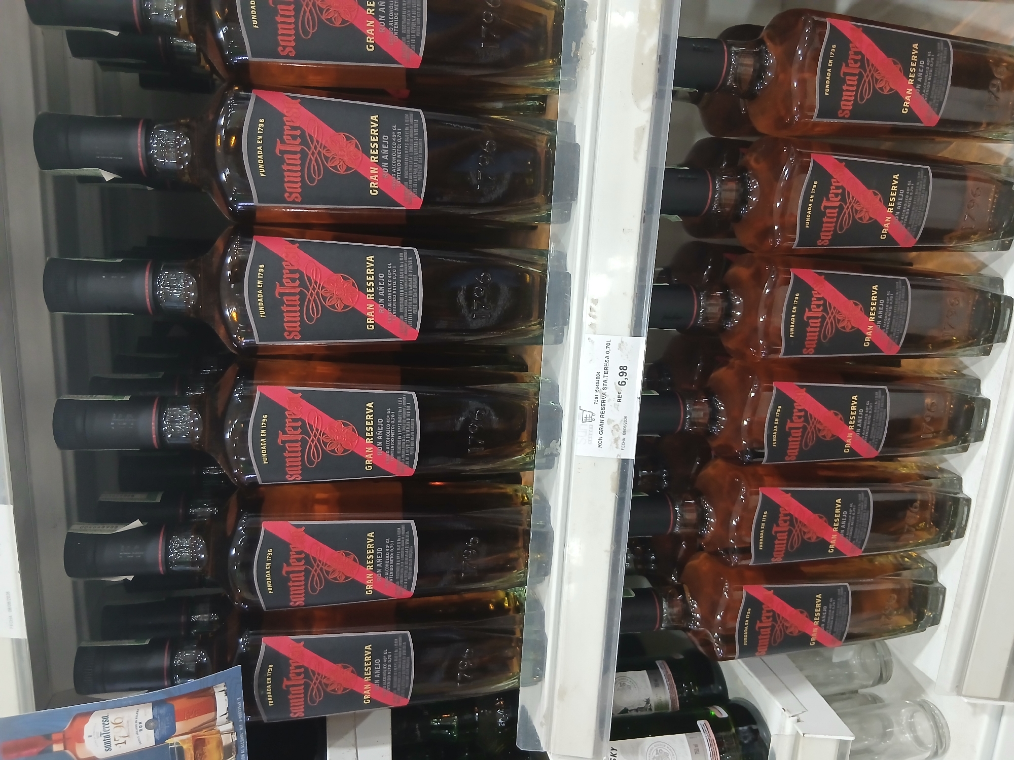 The image shows a close-up view of a retail shelf displaying several bottles of Santa Teresa Gran Reserva Ron Anejo. The bottles are amber-colored and feature black labels with red and white text and graphics. The bottles are arranged in multiple rows, with the front row being most prominent. The background consists of a white shelf and a hint of a white wall. A price tag is visible on the shelf in the lower right corner, indicating "RON GRAN RESERVA STA.TERESA 0,70L" and a price of "6,98" with a date of "08/06/2026". The scene appears to be inside a store, likely a supermarket or liquor store, in Juan Griego, Venezuela. No people or specific environmental features are discernible beyond the retail setting.