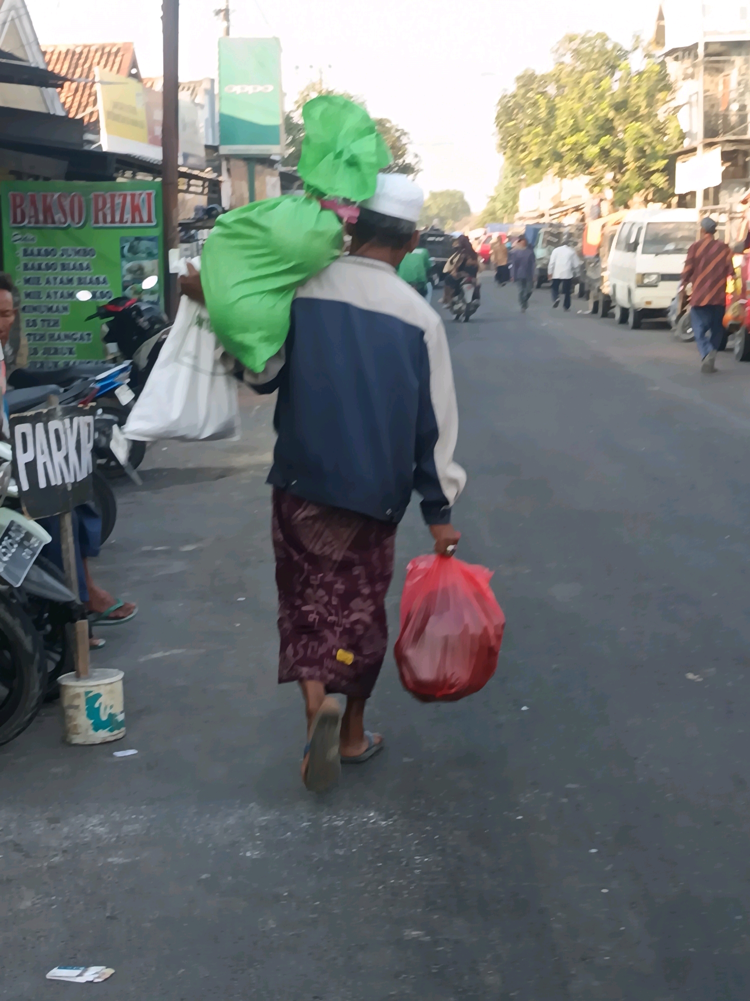 In the city of Blega, Indonesia, a man wearing a white cap, a blue and white jacket, and a patterned sarong walks away from the camera on a paved street. He carries a large bright green plastic bag slung over his right shoulder and a red plastic bag in his right hand. To his left, a white plastic bag is visible. In the background, there are various buildings, including one with a green sign that reads "BAKSO RIZKI" in red lettering. Several vehicles, including motorcycles and a white van, are parked or driving on the street. Trees and utility poles are also present in the environment. The scene appears to be a marketplace or a busy street with shops.