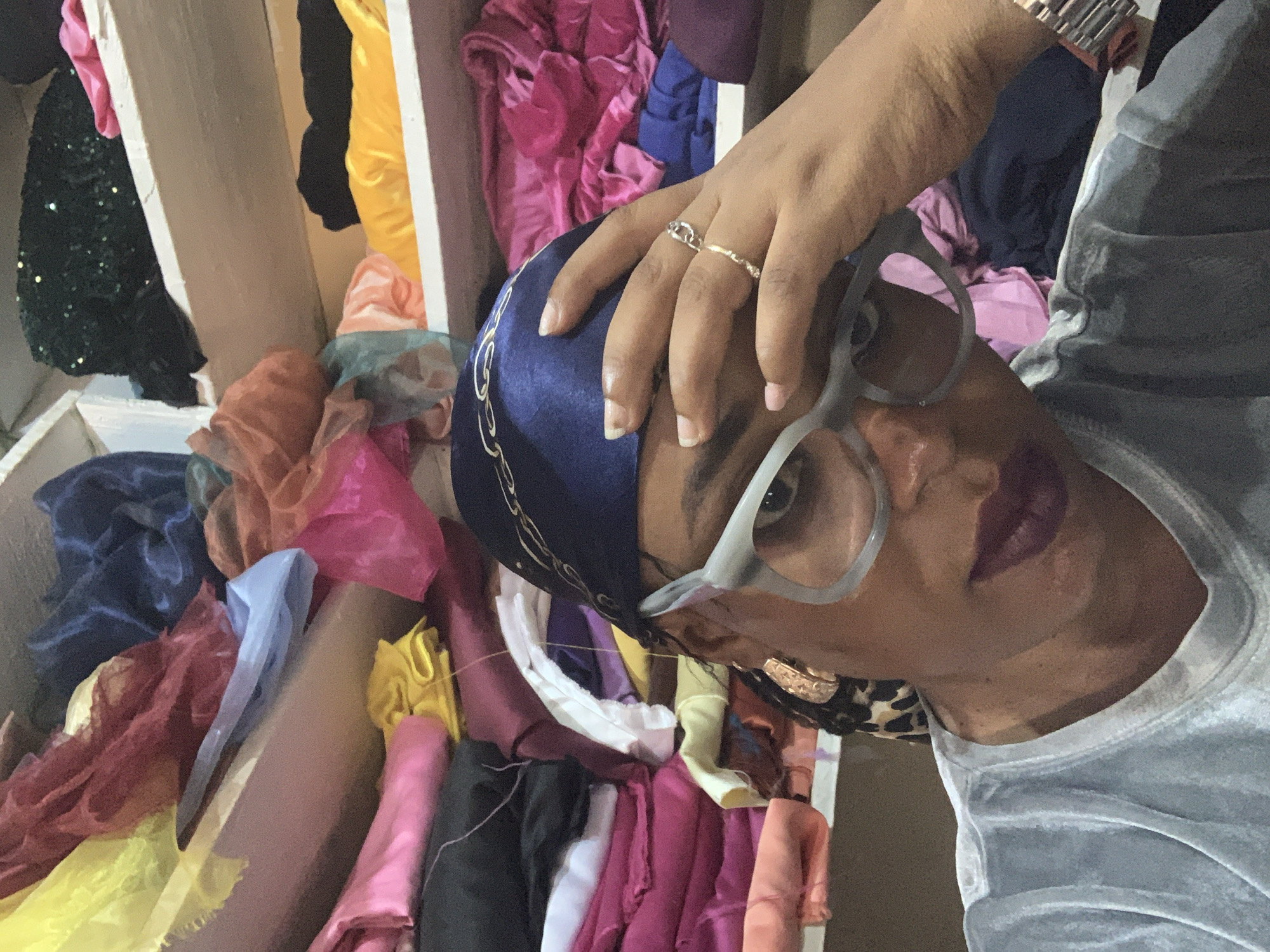 The image shows a person indoors in Sabon Tasha, Nigeria, surrounded by a collection of colorful fabrics. The person is wearing glasses, a blue head covering, and a silver-toned watch, with rings on their fingers. They appear to be touching their head. The fabrics are arranged in a box or shelf.