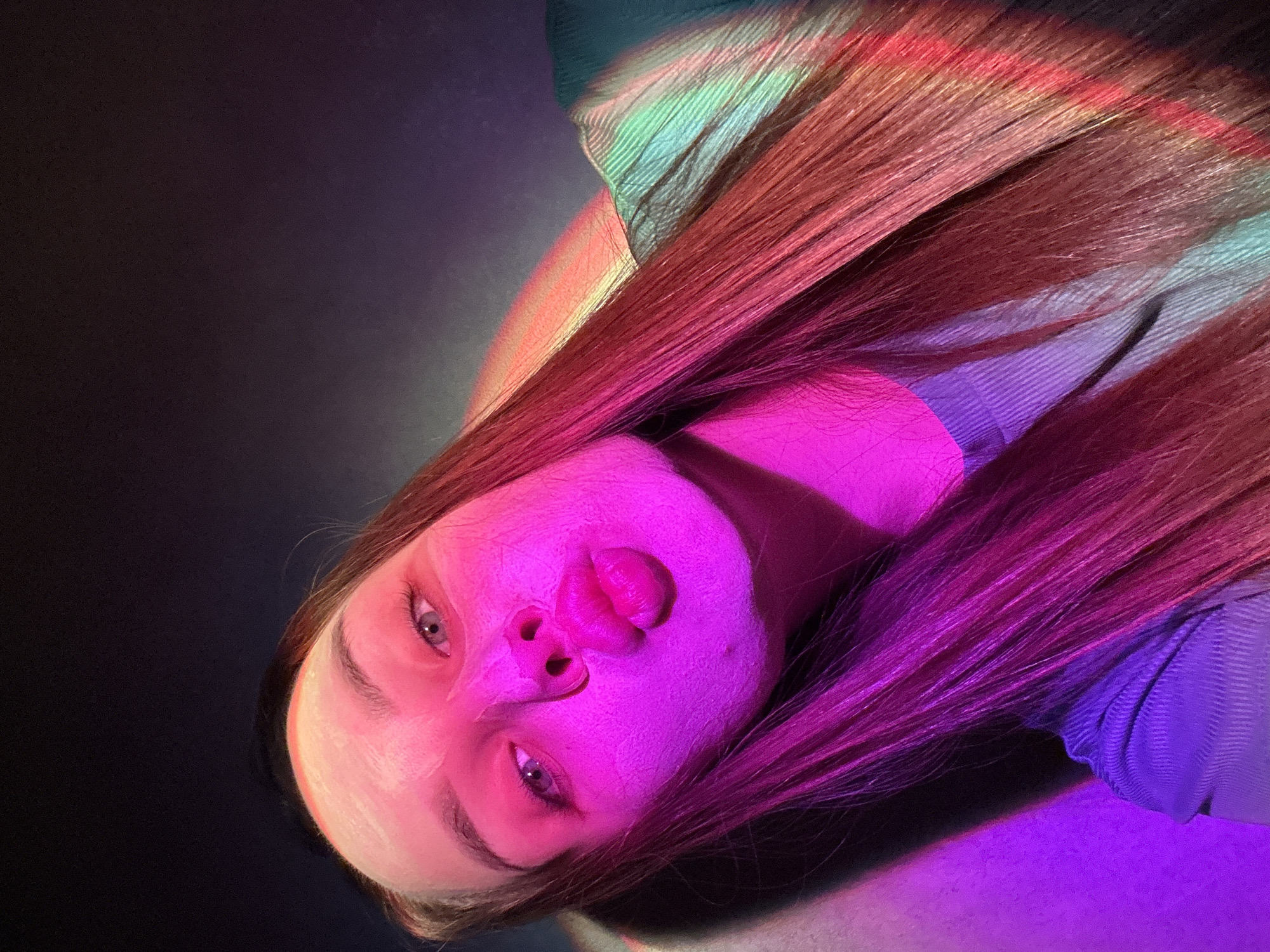 A portrait-style photograph captures a young woman's face and upper body, dramatically lit with vibrant colored lights. The image is oriented with her head tilted to the left, appearing to look upwards if rotated upright. Her long, reddish-brown hair cascades around her, illuminated by distinct streaks of pink, purple, red, and hints of green and blue light. Her face is predominantly bathed in a bright magenta and pink glow on the right side, transitioning to a softer orange-pink and then white on her forehead. She has her lips slightly parted in a subtle pout and makes direct eye contact with the viewer, conveying a somewhat contemplative or alluring expression. Her visible clothing, likely a ribbed top or sweater, appears in shades of teal on her upper left shoulder and purple on her right shoulder/chest area. The background is dark, emphasizing the subject and the dramatic, multi-colored lighting, which creates an artistic and ethereal atmosphere. The setting is likely indoors with controlled lighting, possibly for a photoshoot. There is no visible text in the image. This photo was taken in Chaikovsky, Russia.