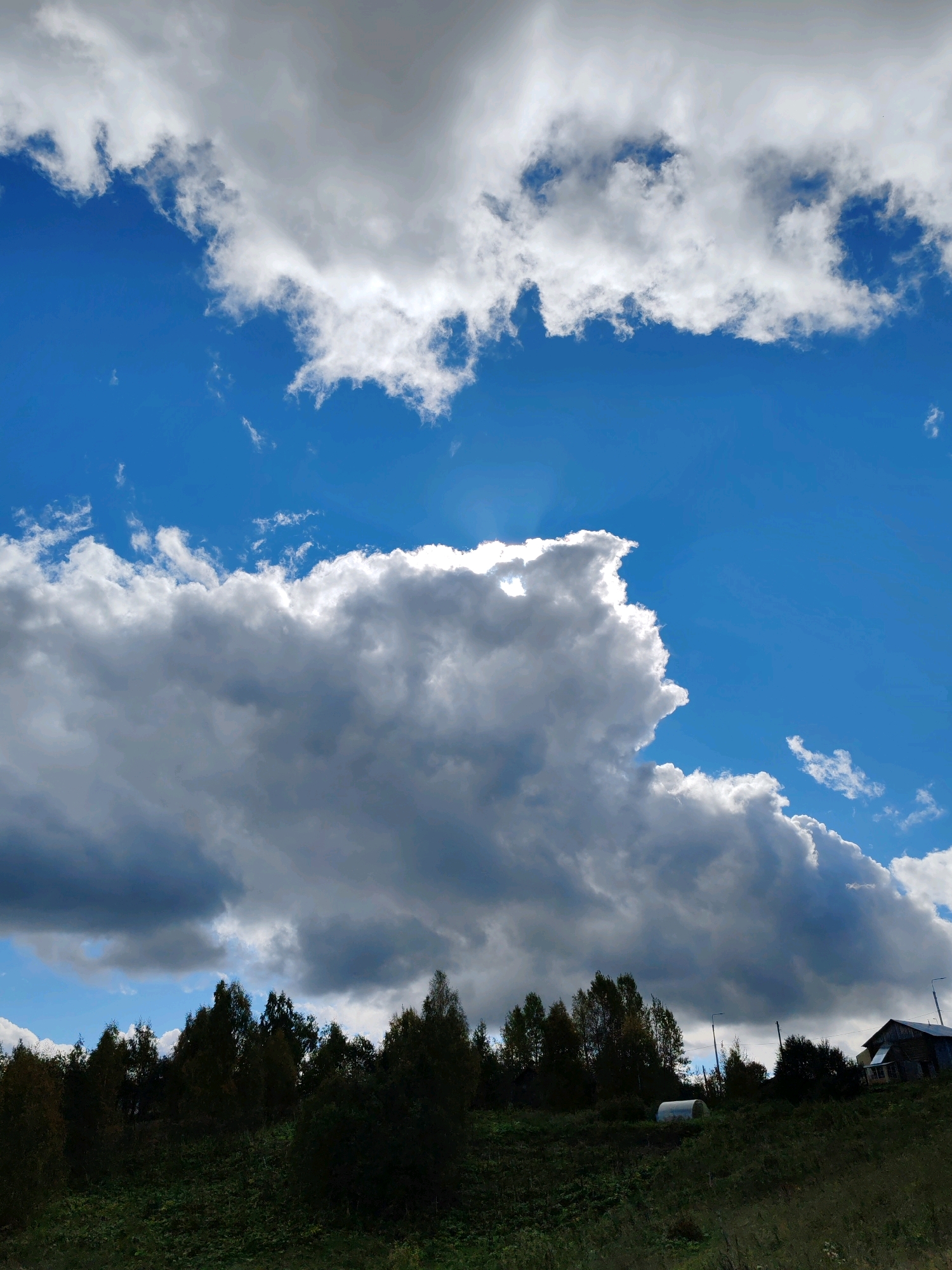 The image depicts a vast, blue sky filled with dramatic, white and grey clouds. Sunlight pierces through some of the clouds, creating bright highlights. Below the sky, a dense line of dark green trees forms a horizon. In the foreground, a grassy hill slopes downwards. On the right side of the hill, a weathered, dark wooden house with a sloped roof is visible. A small, white, arched structure, possibly a greenhouse, is situated on the left side of the hill amidst the greenery. The overall scene suggests a rural or remote location. The provided context indicates the location is Чухлэм, Russia.