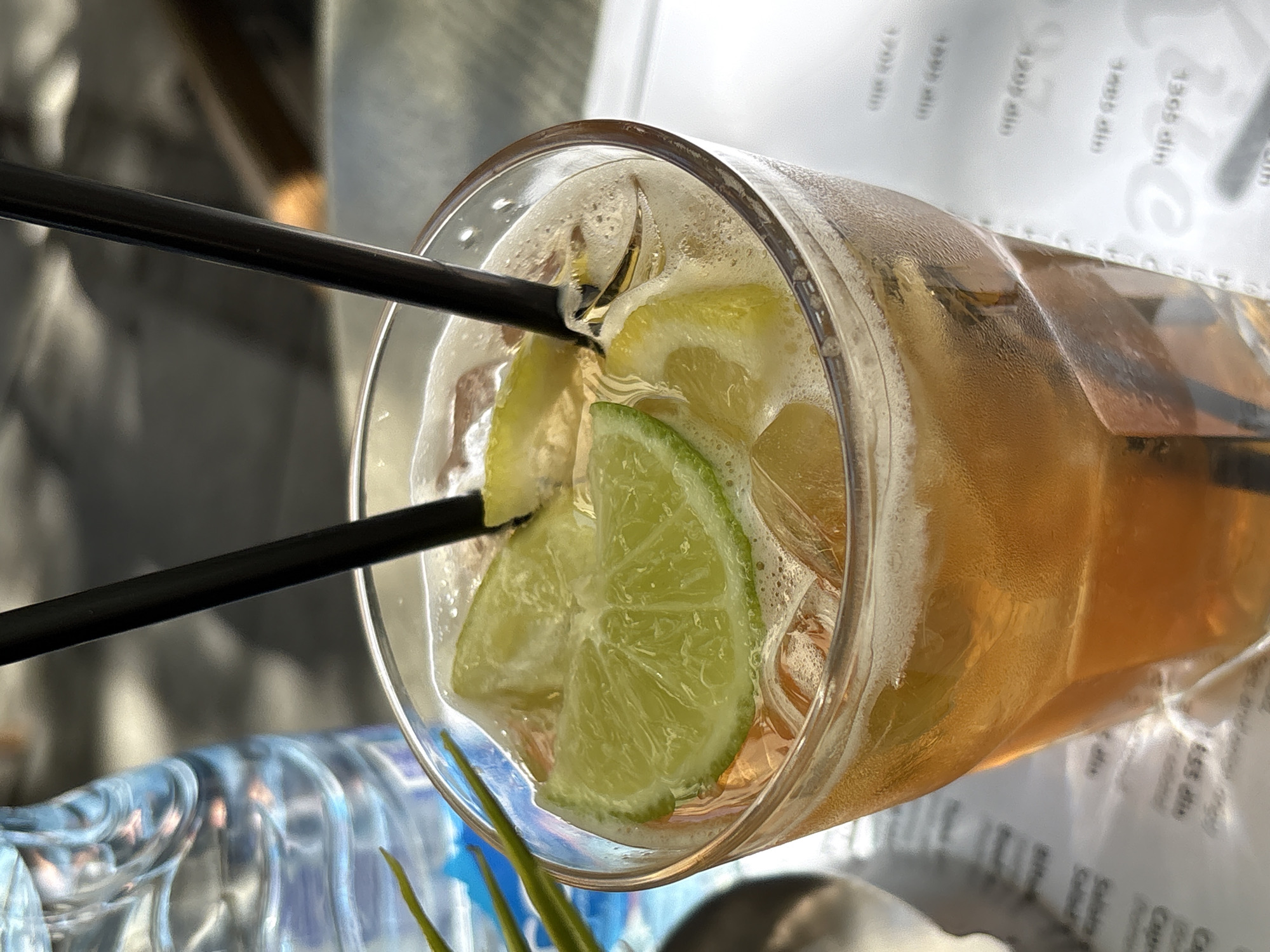 The image captures a close-up view of a tall glass filled with an amber-colored beverage, likely iced tea or a similar drink, adorned with ice cubes and two slices of lime. Two black straws are inserted into the drink. In the background, a partially visible bottle of water with a blue label is present, suggesting an outdoor setting, possibly a cafe or restaurant. The table surface appears to be covered with a white paper mat with some text. The overall lighting suggests it is daytime, and the environment has a casual, refreshing atmosphere. The context of Belgrade, Serbia, is not directly ascertainable from the visual elements alone, as no specific landmarks or cultural indicators are present in the frame.