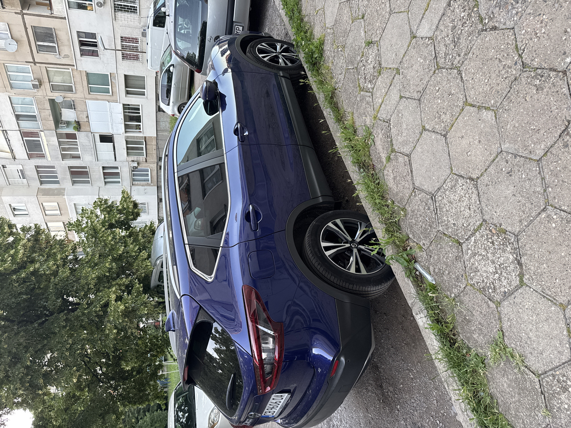 A dark blue Nissan Qashqai is parked on the side of a street in Sofia, Bulgaria, next to a concrete hexagonal paved sidewalk with grass growing in the cracks. The car is positioned in front of a multi-story residential building with numerous windows and balconies. In the background, other parked vehicles are visible, including a silver car and a white van. The scene appears to be a typical urban residential area.