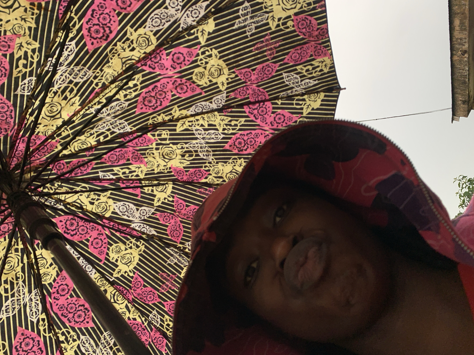 The image presents a low-angle perspective looking upwards, featuring a person holding an umbrella. The primary subject is a person with dark skin, likely a child or young adult, whose face is partially visible in the lower right. They are wearing a hooded garment, possibly a jacket, which appears dark with a reddish or pink patterned lining. Their lips are pursed in a playful or posed expression.
Dominating the left and upper portions of the frame is the underside of an open umbrella, showcasing a distinct and colorful pattern. The umbrella's design features a black background with thin, horizontal yellow stripes. Overlaid on these stripes are bold floral and leafy motifs in alternating pink and yellow, with some white elements also present. The internal ribs and central shaft of the umbrella are clearly visible, fanning out across the patterned fabric.
In the background, visible in the upper right, is a bright, overcast sky. A section of a weathered building or structure, possibly with a corrugated roof, is also present. A thin, dark wire stretches horizontally across the sky, connecting towards the building. A small cluster of green foliage is barely visible on the far right edge. The scene appears to be set during daytime in Rafin Par, Nigeria. No visible text is present in the image.