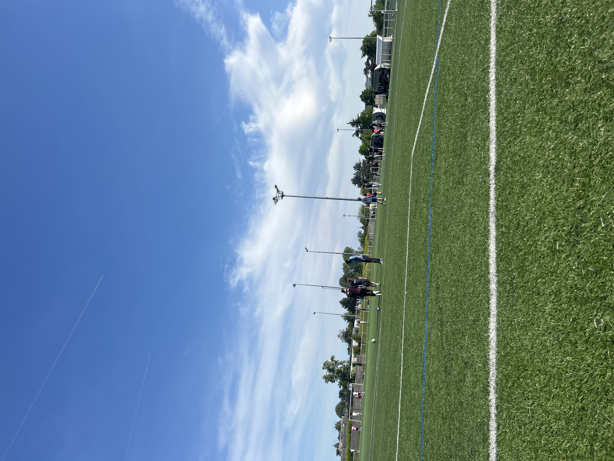The image depicts a sunny day at a soccer field in Champigny-sur-Marne, France. A group of individuals are present on the field, seemingly engaged in practice. The environment includes the soccer field, marked with white and blue lines, stadium lights, and surrounding buildings. The sky is clear with some scattered clouds. Two jet trails are visible.