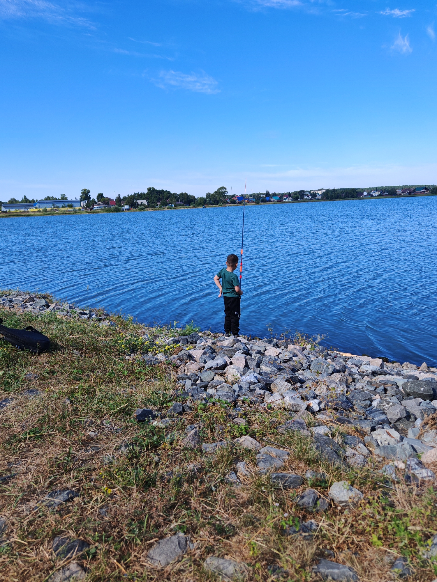A young boy stands on a rocky shore, holding a fishing rod. He is wearing a green t-shirt and black pants. The boy is looking out at a large body of blue water, which reflects the clear blue sky above. In the distance, across the water, is a shoreline with houses and trees. The foreground is covered in dry grass and rocks, with some yellow flowers growing among the rocks. A black bag lies on the grass to the left of the boy. The scene appears to be in Chernakovo, Russia.