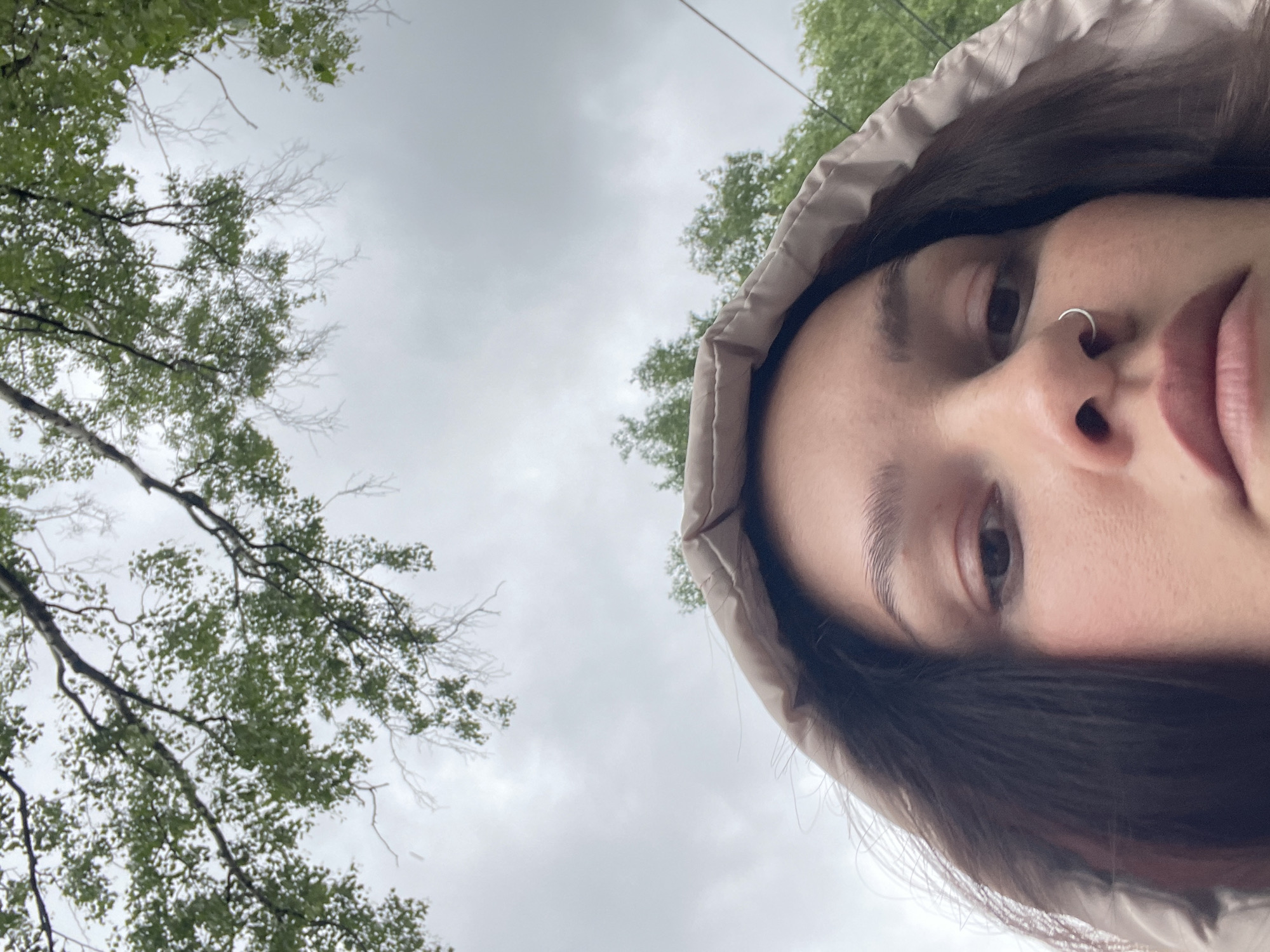 A low-angle selfie shows a woman with a nose ring, wearing a hooded jacket. Above and to the left of her is a view of tree branches with green leaves against an overcast sky. The location is presumed to be in Izhevsk, Russia.