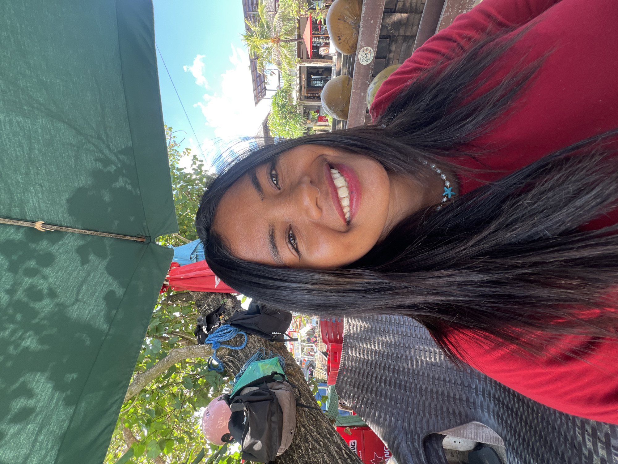 A young woman with long, dark hair and a red shirt smiles at the camera. She is wearing a pearl necklace with a blue starfish pendant. The woman is seated in a woven chair, likely outdoors, under a green umbrella, with dappled sunlight filtering through leaves. Behind her, a tree is visible with several backpacks and a pink helmet hanging from its branches. In the background, there are buildings, palm trees, and large, round objects that appear to be coconuts. The setting suggests a tropical location, possibly a beachside cafe or restaurant in Kuta, Indonesia, given the architectural style and vegetation. The sky is bright blue with scattered clouds.