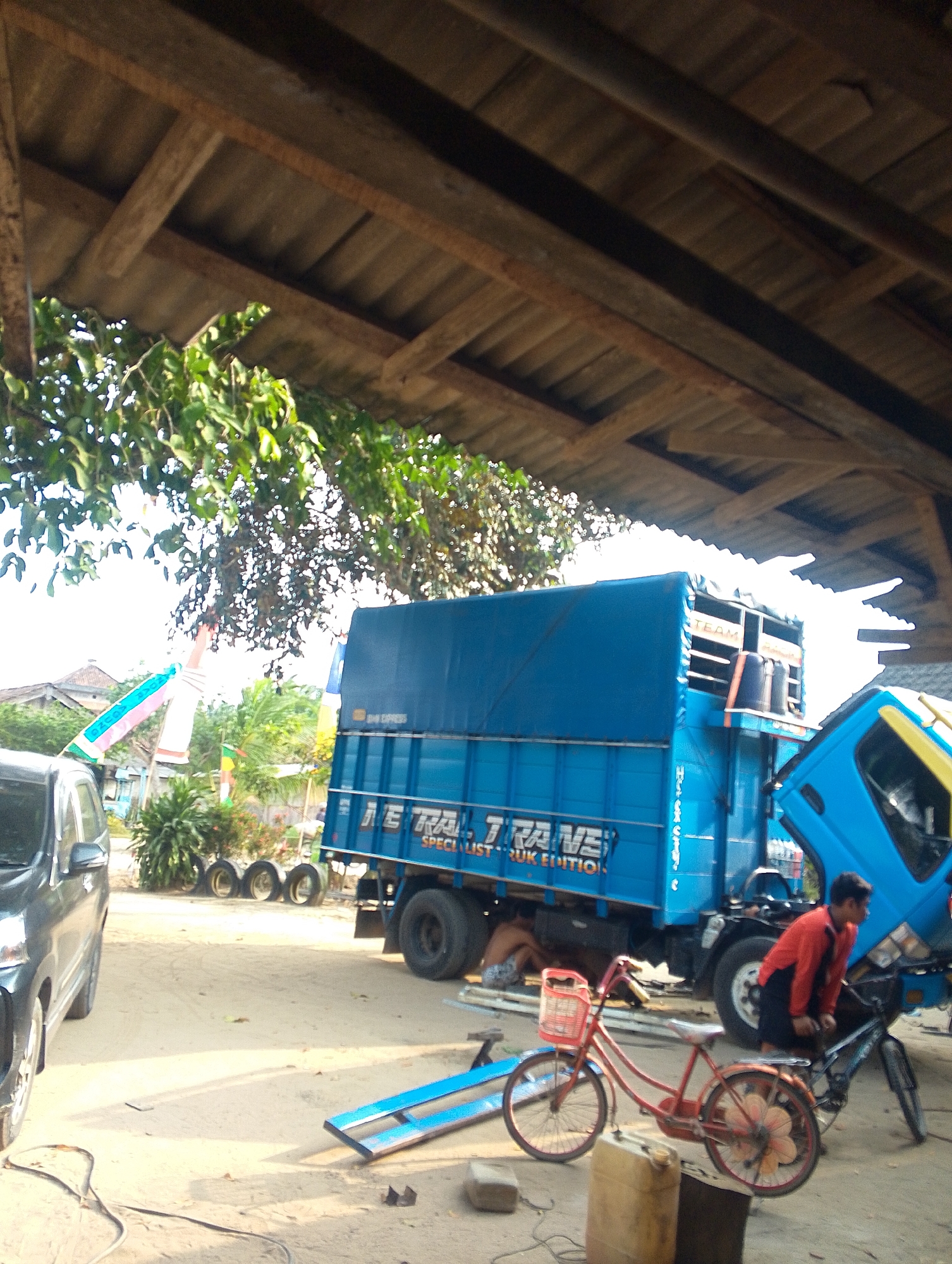 Underneath a rustic, wooden-raftered roof, a scene unfolds in what appears to be Karang Mulyo, Indonesia. A large blue truck with the text "J.I.T. TRANS" and "SPECIALIST TRUK EDITION" emblazoned on its side is parked on a sandy lot. The truck's hood is open, revealing mechanical components. A man in a red jacket is leaning over the truck, appearing to work on it. In the foreground, a red bicycle with a basket is parked. Several other bicycles and spare tires are visible nearby. A dark-colored car is parked to the left of the frame, and a yellow jerrycan sits on the ground. The environment is dusty and appears to be an outdoor workshop or parking area, with some greenery and buildings visible in the background.