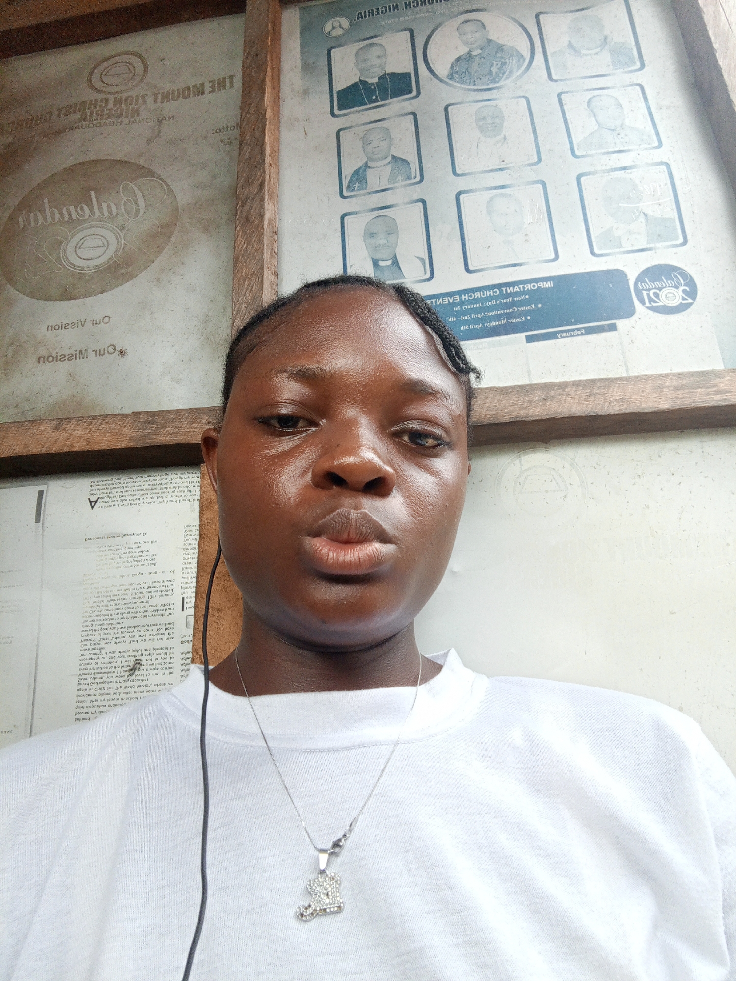 A young person, appearing female, is captured in a close-up selfie or portrait, looking directly at the camera. Their dark skin is illuminated by bright lighting, highlighting a slightly shiny complexion. They have braided hair pulled back and wear a plain white t-shirt. A silver necklace with a sparkling, intricate pendant hangs around their neck, and a black wired earphone is visible near their face. Their expression is neutral, with lips slightly pursed in a posed manner.
The setting appears to be an indoor or covered area, possibly in Mbribit, Nigeria, with two large, worn information boards serving as the backdrop, separated by a rough wooden beam.
The board on the left, partially obscured by the subject, features reversed text and a logo. Visible reversed phrases include "Our Vision" and "Our Mission," and a larger, reversed "THE MOUTH SION CHURCH NIGERIA" (possibly "Zion") is discernible. A prominent reversed circular logo, possibly for "Calabana," is also present. This board also contains dense, largely illegible reversed text.
The board on the right displays a grid of nine framed black and white portrait photographs of men, many appearing to be in clerical or official attire. Below these images, reversed text reads "IMPORTANT CHURCH EVENTS" and "2021 Calendar," suggesting it's an organizational bulletin board. Both boards show signs of dirt, wear, and possibly water damage, with some text difficult to read due to reflections and grime. The overall impression is a casual portrait taken against a backdrop of faded institutional information.