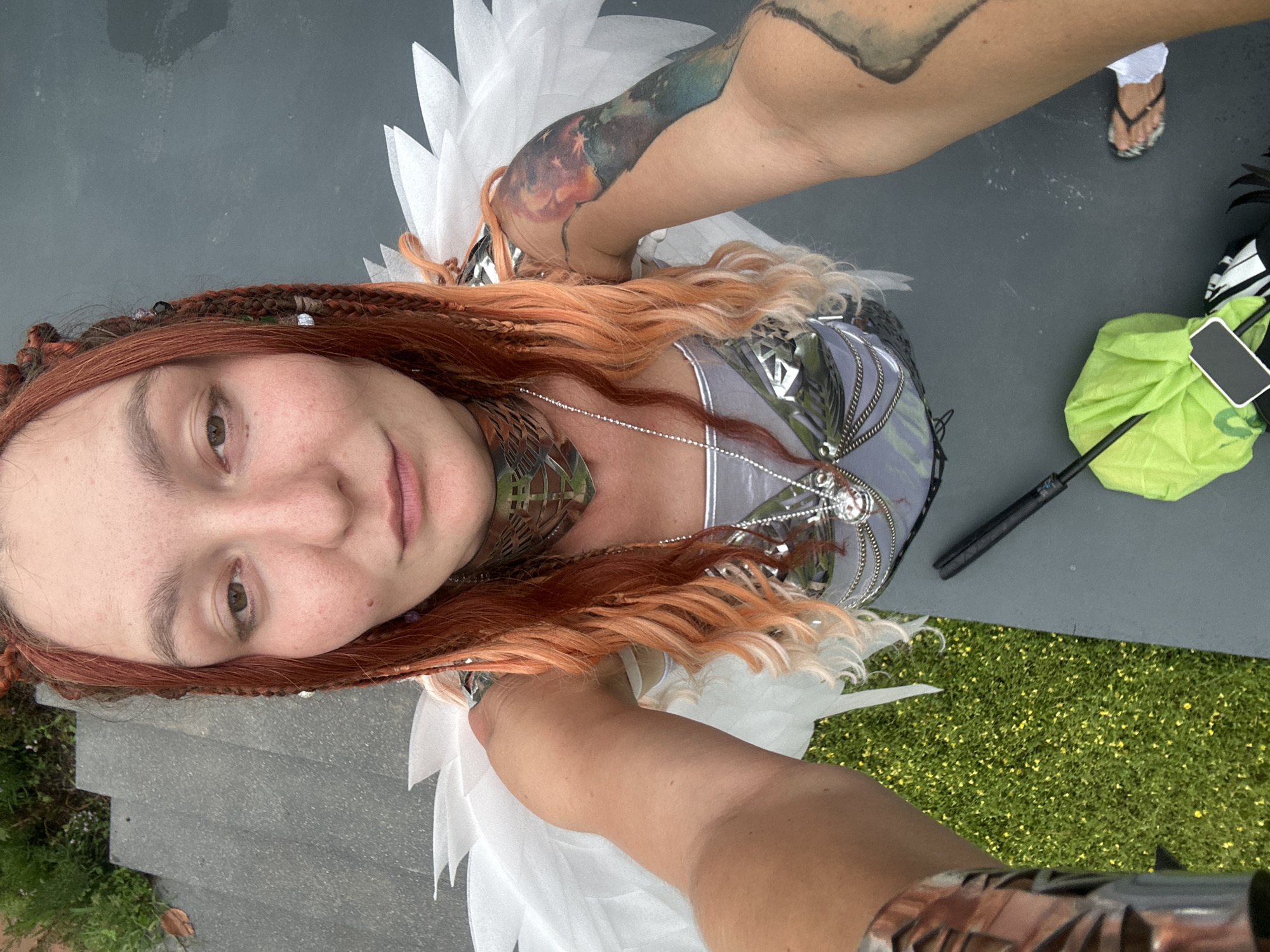 A woman with auburn braided hair and streaks of blonde is taking a selfie outdoors. She is wearing a silver, metallic-looking outfit with white feather-like wings attached to her shoulders. Around her neck is a large, ornate silver collar. She has visible tattoos on her right arm. In the foreground, a black object, possibly a staff or umbrella, rests on a dark gray surface next to a bright green bag. To the left, there is green grass with yellow flowers. Behind her, gray steps are visible with some foliage at the top. The lighting suggests it is daytime.