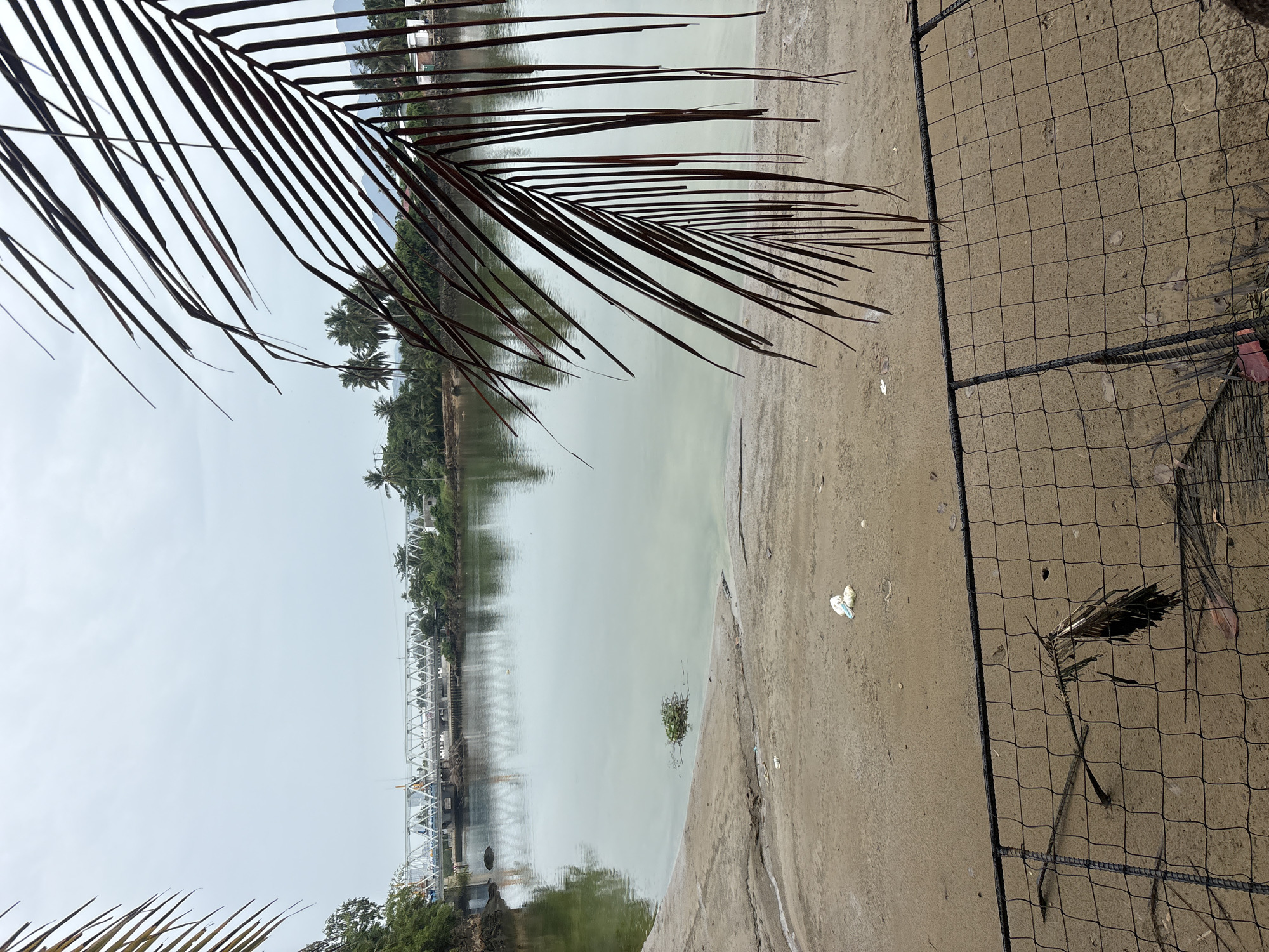A wide shot captures a tranquil river scene in Nha Trang, Vietnam, with a light gray, overcast sky. In the foreground, a rough, sandy embankment leads down to the murky, greenish water. A black metal mesh fence, partially obscured by palm fronds and debris, runs across the bottom of the frame. Scattered white plastic trash is visible on the sand. The river is spanned by a long, white truss bridge with vehicles crossing it, indicating it's a functional transportation route. Lush green trees and vegetation line the opposite bank, with some buildings and palm trees visible further inland. In the distance, mountains are faintly discernible. The image composition is framed by dark brown palm fronds in the upper right and left corners, creating a natural border.