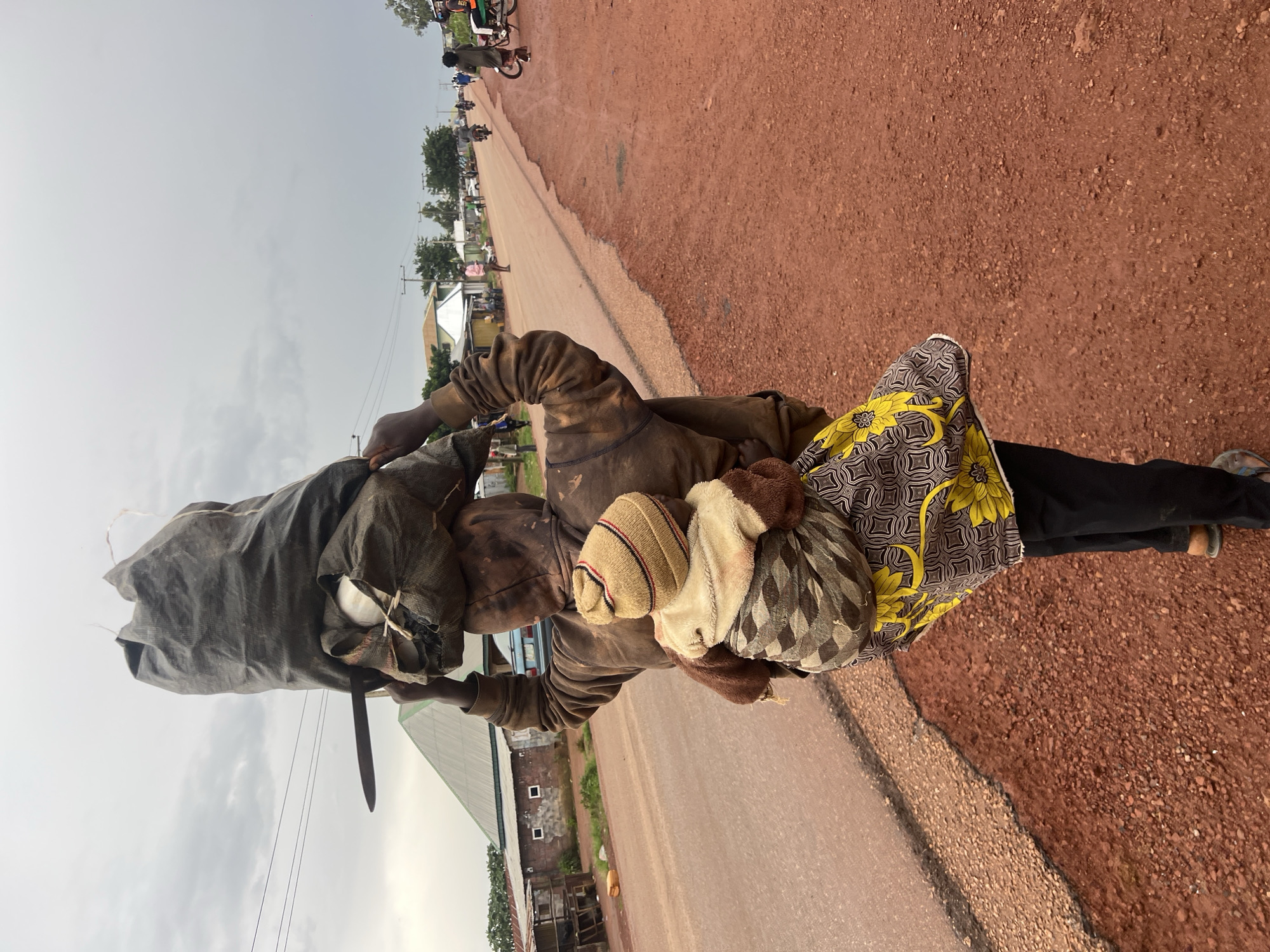 A person walks on a dusty, red dirt road on the outskirts of Durumi, Nigeria, carrying a large sack of goods balanced on their head and a baby wrapped to their back. The person is wearing a brown hooded sweatshirt and black pants. The baby is wearing a striped knit hat and a beige and brown blanket. The road is lined with small buildings and trees. In the distance, several motorcycles and a bicycle can be seen. The sky is overcast.