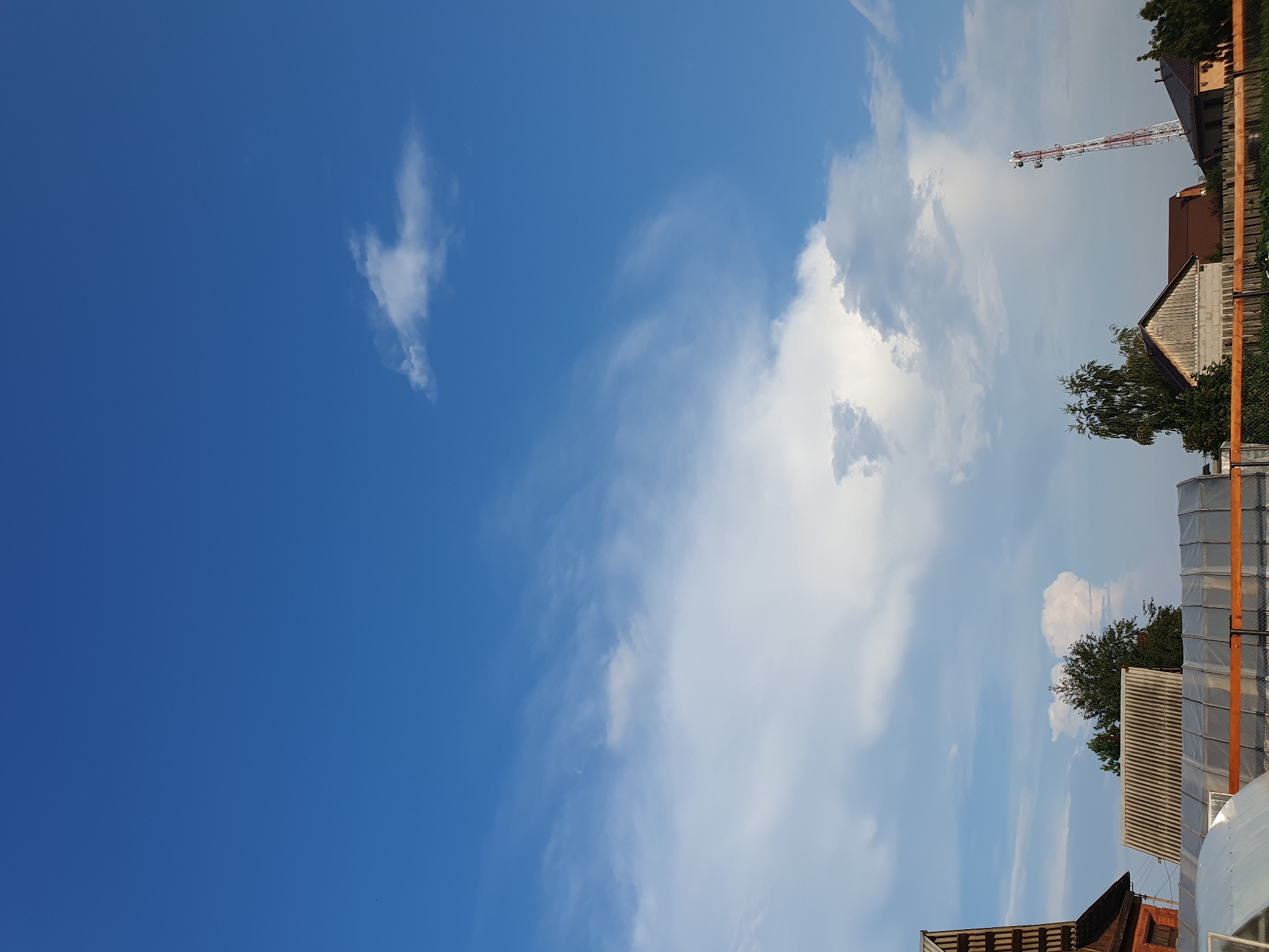 The image shows a rural scene in the city of Meget, Russia, under a bright blue sky with scattered clouds. In the foreground, there are several structures, including a greenhouse with a white tarp, a building with a corrugated roof, and a brick building with a wooden roof. Further back, there are trees and houses. A tall telecommunications tower with red and white stripes stands prominently in the middle ground. The scene appears to be a residential area in the countryside.