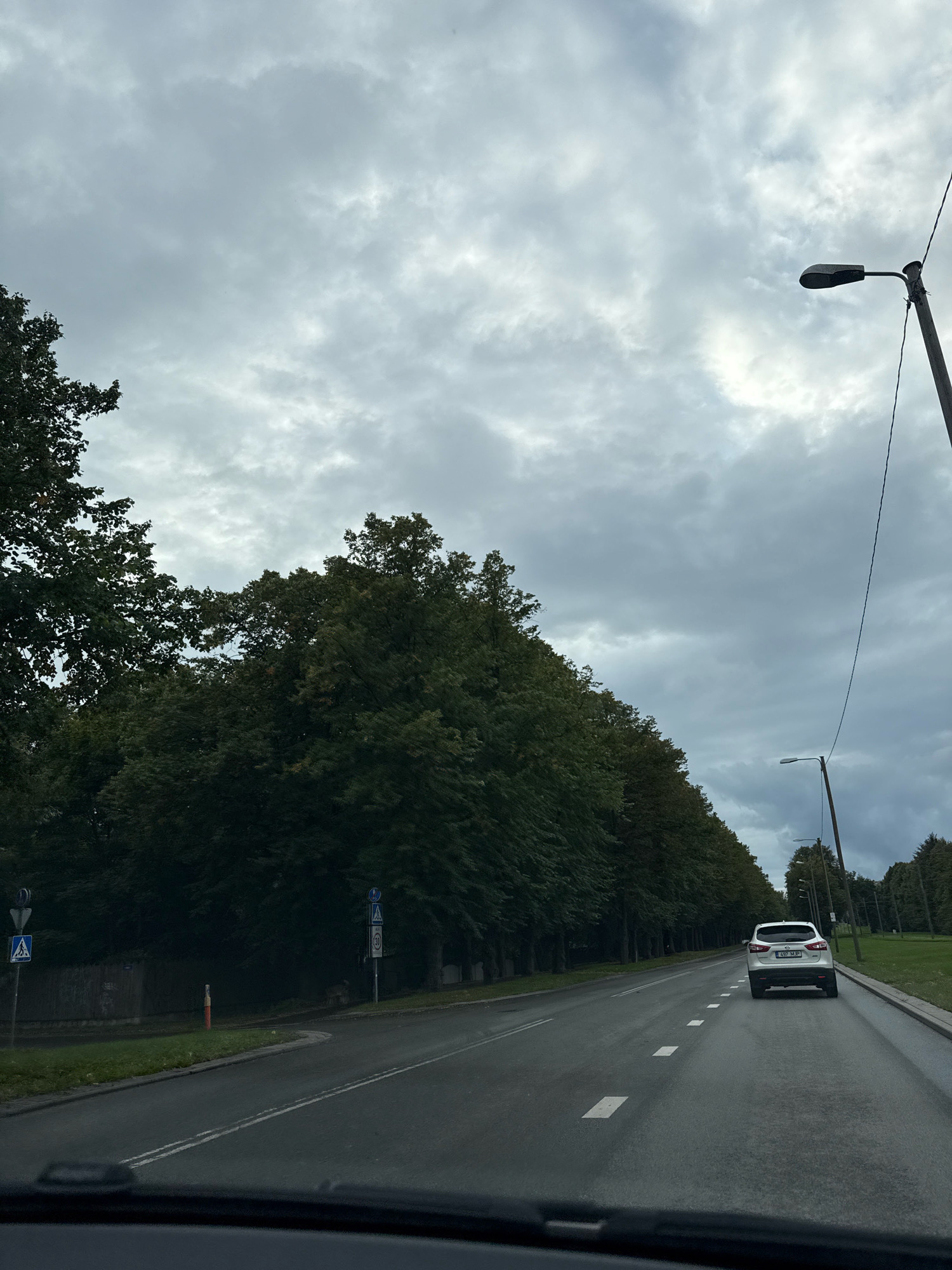 An exterior, eye-level, medium shot captures a street scene in Tallinn, Estonia, under an overcast, cloudy sky. The perspective is from inside a vehicle, looking forward through the windshield. A white SUV is driving away from the camera on the right lane of a wet asphalt road. The road is lined with tall trees with dark green foliage on the left and a grassy area with more trees on the right. There are streetlights along the road. Traffic signs are visible on the left side of the road, including a blue square sign with a white pedestrian symbol and a circular blue sign with a white arrow. A red and orange bollard stands near the side of the road on the left. The overall impression is a somber, somewhat dreary atmosphere due to the weather.