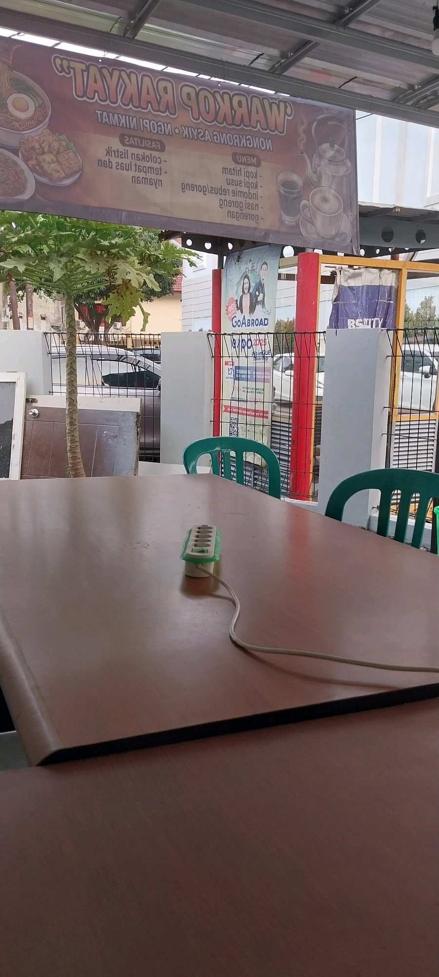 An outdoor seating area of a roadside food stall is depicted in South Tangerang, Indonesia. A wooden table occupies the foreground, with a white power strip featuring six outlets placed centrally on its surface. Green plastic chairs are visible in the background, hinting at customer seating. Behind the chairs, a black metal fence separates the seating area from the street, where cars are parked. A banner advertising "WARKOP RAKYAT" hangs above the stall, featuring images of food and drinks. To the right of the banner, a poster for "GOABROAD" with two individuals is displayed on a red pillar. The overall scene suggests a casual dining establishment in an urban setting.