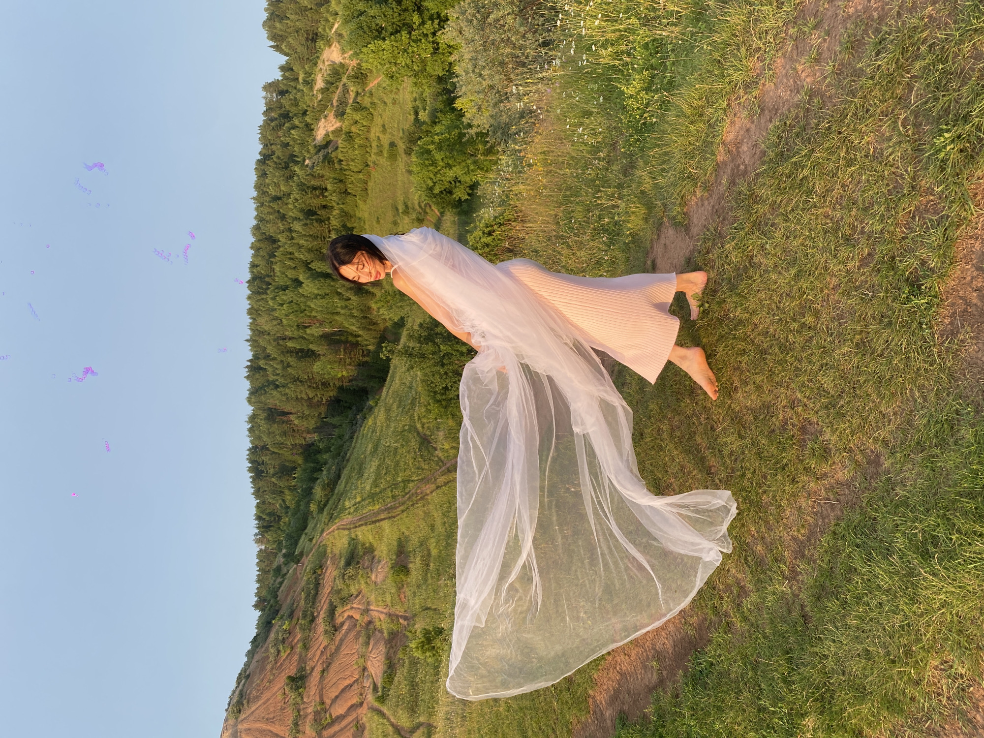 An adult female is standing barefoot on a grassy hillside, holding a sheer, white veil that billows out to her left. She is wearing a light pink, ribbed maxi dress and has dark hair and glasses. The background features a sparsely vegetated, reddish-brown hill to the left, a dense green forest line across the middle distance, and a clear blue sky above. A dirt path winds up the grassy slope. There are some blurred purple shapes in the sky. The location is the city of Olkhovka, Russia.
