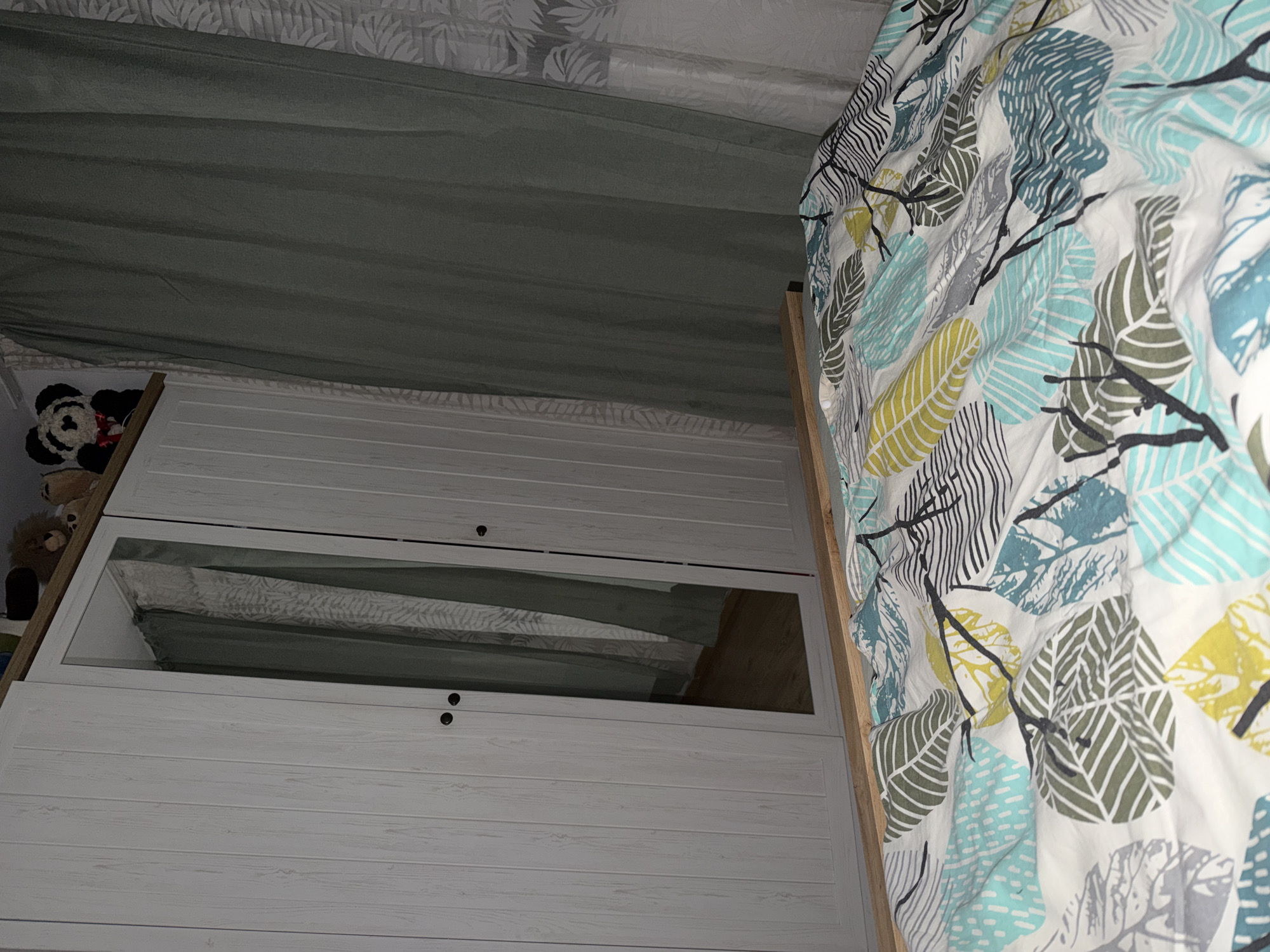 The image displays an indoor scene, likely a bedroom, presented from an upward-looking perspective. On the right, a bed is visible, adorned with a white duvet featuring a decorative pattern of stylized leaves in shades of blue, green, yellow, grey, and black. To the left, a white wooden wardrobe or cabinet stands, constructed with horizontal paneling. The upper section of the wardrobe has a small, round black handle. Below this, a rectangular mirrored or glass panel is incorporated, reflecting a light grey-green curtain or blind and a section of the ceiling above it, which displays a subtle leaf pattern. Another small, round black handle is positioned beneath the mirrored panel. Above the wardrobe, a light grey-green curtain or fabric appears gently folded. In the upper-left corner, behind the wardrobe, partially visible soft toys include a brown stuffed animal and a black and white floral object. A light brown wooden frame runs along the right side of the wardrobe. The room is dimly lit, suggesting an interior setting with limited natural light or artificial illumination. No people or activities are present, and no text is visible. The scene does not contain any specific visual cues to its location in Plovdiv, Bulgaria, beyond being a typical indoor living space.