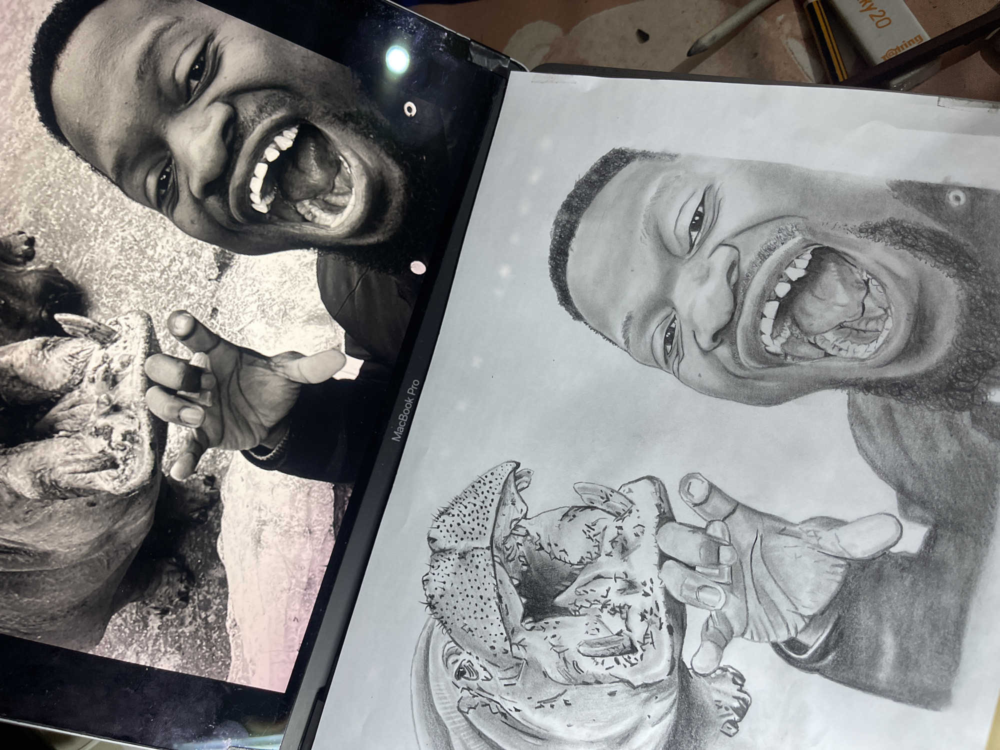 The image, depicting an indoor setting understood to be within Jos, Nigeria, presents a MacBook Pro laptop displaying a black and white photograph positioned directly above a detailed graphite pencil drawing on white paper. The laptop screen shows a dark-skinned male with a short beard, wide-eyed and laughing with his mouth open, revealing several missing upper front teeth. His left hand, with slightly curled fingers, is extended towards the similarly monochromatic, wide-open mouth of a hippopotamus visible to the upper left. The pencil drawing meticulously replicates this scene, mirroring the man's expressive face, his interaction with the hippo's maw, and the texture of both subjects. To the right of the drawing, an eraser marked "rOtring KY20" and what appears to be a blending stump or pencil are visible on the underlying surface.