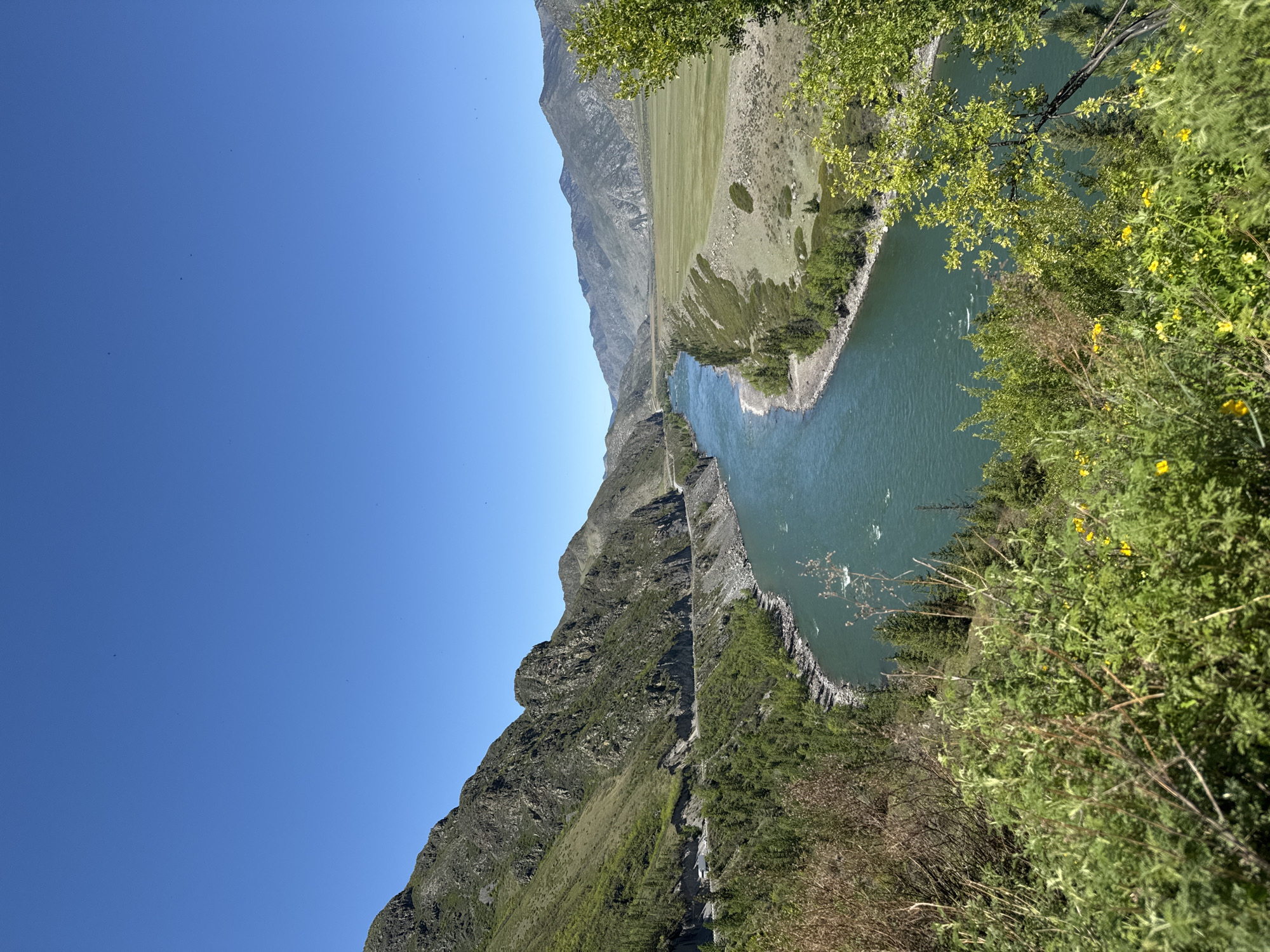 A wide-angle landscape shot shows a turquoise river winding between mountains under a clear blue sky in Malyi Yaloman, Russia. A rugged, steep mountain covered with vegetation rises on the left side of the image, while a flatter, grassy area with some rocky patches extends on the right side. Lush green trees and bushes fill the foreground on the right, partially obscuring the river. A road can be seen carved into the side of the mountain.