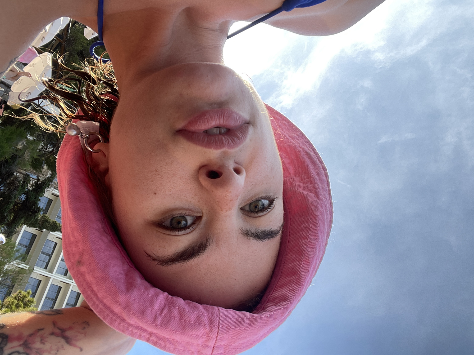 A selfie portrait of a woman with fair skin and green eyes, wearing a pink hooded sweatshirt, taken from a low angle. She is facing the camera with a slight upward tilt. Her lips are slightly parted. She is wearing a blue bikini top. Her hair is dark, with some strands appearing wet or damp. She has a tattoo visible on her shoulder. A silver earring is visible in her left ear.
The background shows a sky with scattered clouds. The presence of buildings, trees, and umbrellas suggests an outdoor setting in a populated area. Specifically, the buildings are consistent with structures in Rafailovići, Montenegro.