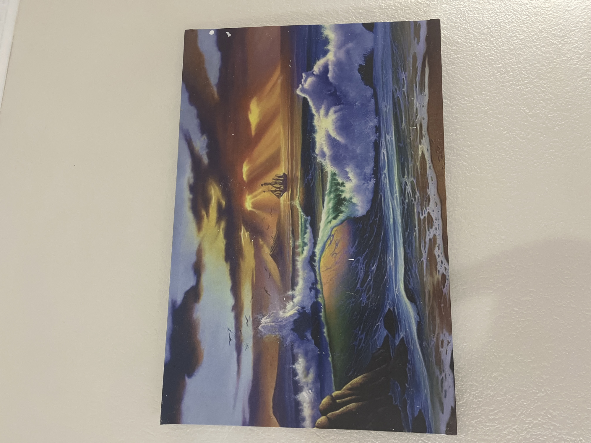 The image shows a painting of a seascape at sunset. A tall ship with sails is visible in the distance, sailing towards the sun. Waves are crashing on a rocky shore in the foreground. The sky is filled with dramatic clouds, lit by the setting sun. The painting is hanging on a textured, off-white wall. There are no people visible in the painting or in the environment. The location in the city of Bashkia Durrës, Albania is not depicted in the image.
