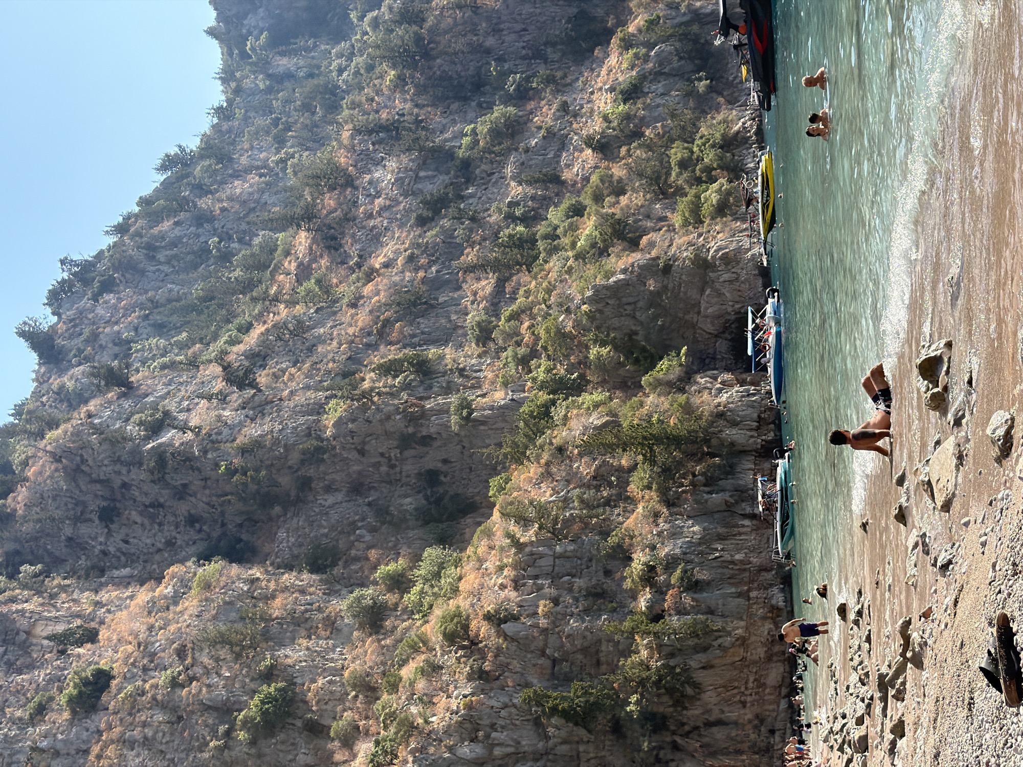 This image depicts a scenic view of Butterfly Valley Beach, located in Fethiye, Turkey. The beach is characterized by its pebbly shore, with clear, turquoise waters lapping at the coastline. Towering, rugged cliffs covered in sparse vegetation dominate the background, creating a dramatic natural amphitheater. Several boats, including speedboats and a yellow personal watercraft, are anchored in the bay, suggesting a popular tourist destination.
On the beach, a scattering of individuals are engaged in various beach activities. In the foreground, a man is seated on the shore, facing the water. Further down the beach, people are seen wading and swimming in the sea. A group of people are gathered at the edge of the water on the left side of the frame. The overall atmosphere is one of a bustling, sunny day at a natural, picturesque beach.