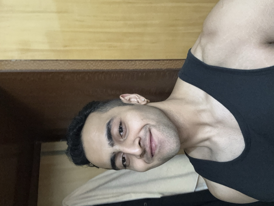 A person wearing a black tank top is lying on a bed. The individual has dark hair and fair skin. Their head is slightly tilted, and they are looking directly at the camera with a slight smile. A gold earring is visible on the left ear. The background includes a wooden headboard.