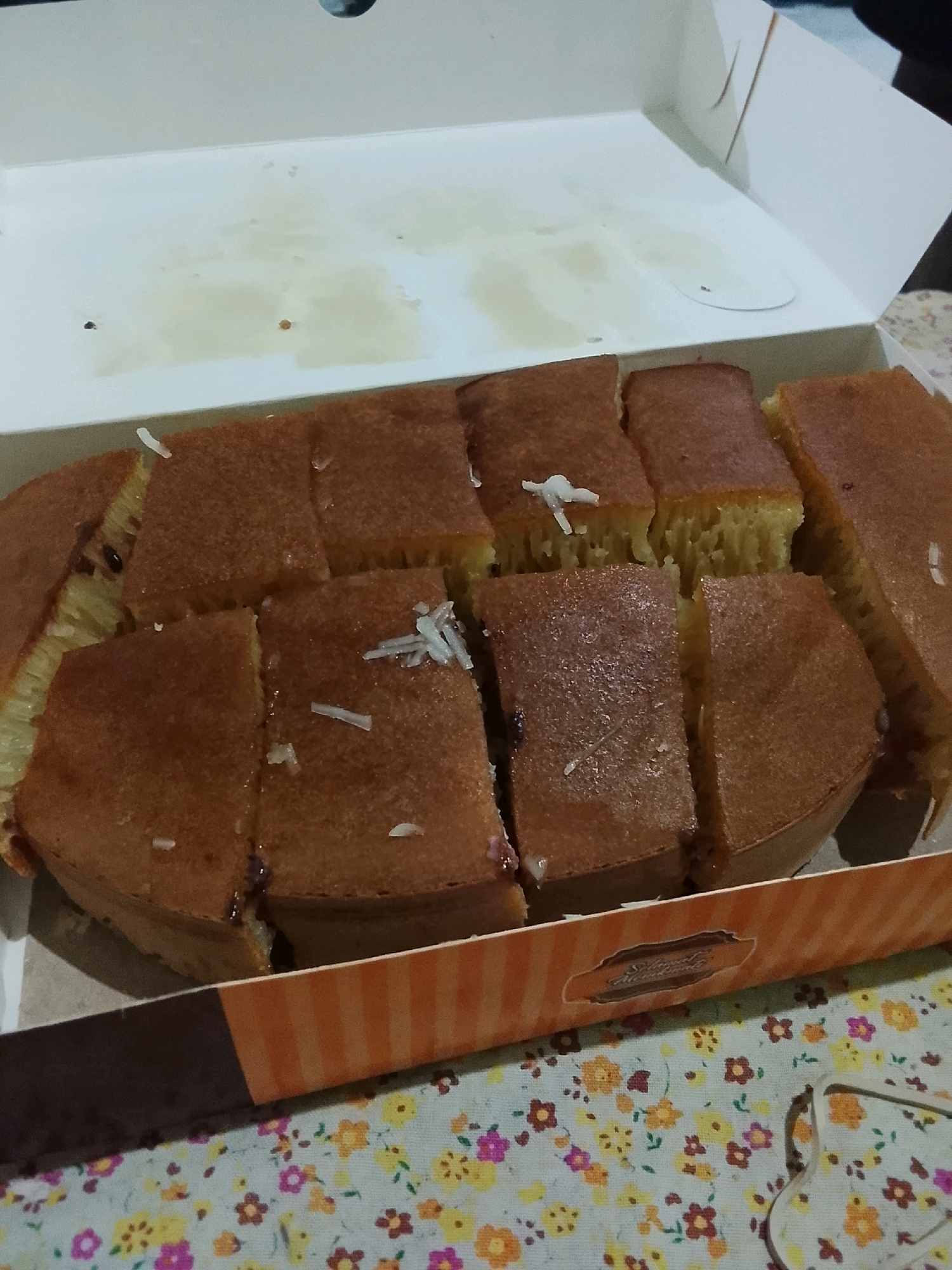 A close-up shot captures a portion of a sweet martabak, a popular Indonesian pancake, presented in a white cardboard box with an orange and brown striped border. The martabak has been sliced into rectangular pieces, revealing its thick, spongy texture and a filling that appears to include chocolate sprinkles and grated cheese. Several pieces are topped with strands of what looks like coconut shavings. The box rests on a floral patterned tablecloth. The scene suggests a casual setting, likely a home or a food stall in Ujungrusi, Indonesia, given the culinary context of the sweet martabak.