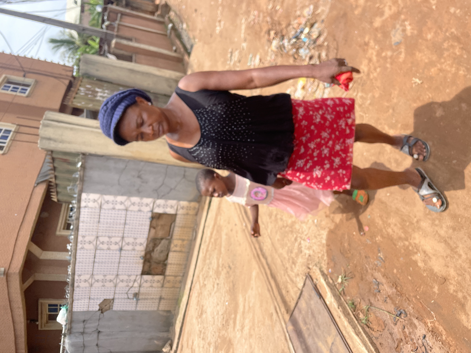 The image, taken in Nkpor, Nigeria, presents a street scene showing an adult woman and a young girl. The woman wears a navy blue knit cap, a black tank top with white detailing, and a red floral skirt. She holds a red object in her outstretched right hand. Her feet are clad in grey and silver sandals. The young girl, partially obscured behind the woman, wears a light pink dress with a round design on the front. The street is unpaved, with patches of debris and standing water. Buildings with cracked walls and partially tiled facades line the side of the street. Overhead, a tangle of electrical wires are visible against the sky.
