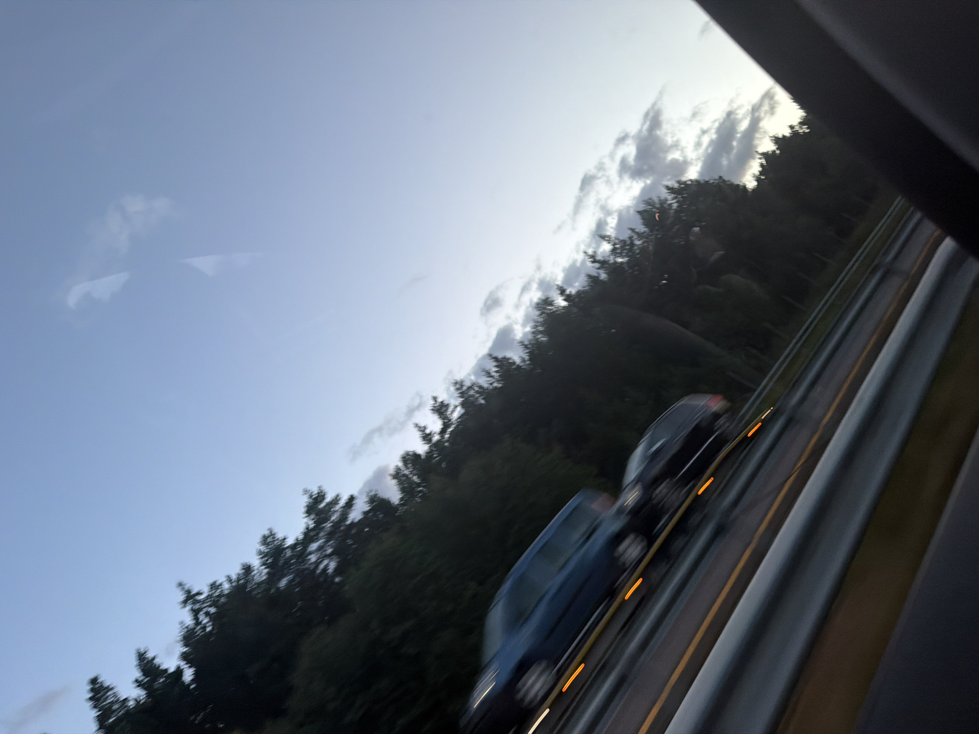 The image depicts a highway scene at dusk or dawn, with a clear sky and scattered clouds. A dark-colored car is in the foreground, moving on the highway. The car is partially obscured by motion blur, but its headlights and taillights are visible as streaks of light. To the left of the highway, a dense line of dark green trees forms a natural barrier. The highway appears to be a multi-lane road with guardrails and yellow lane markers. The overall impression is that of a vehicle traveling at speed on a rural or semi-rural highway in Berg i Østfold, Norway, during twilight hours.
