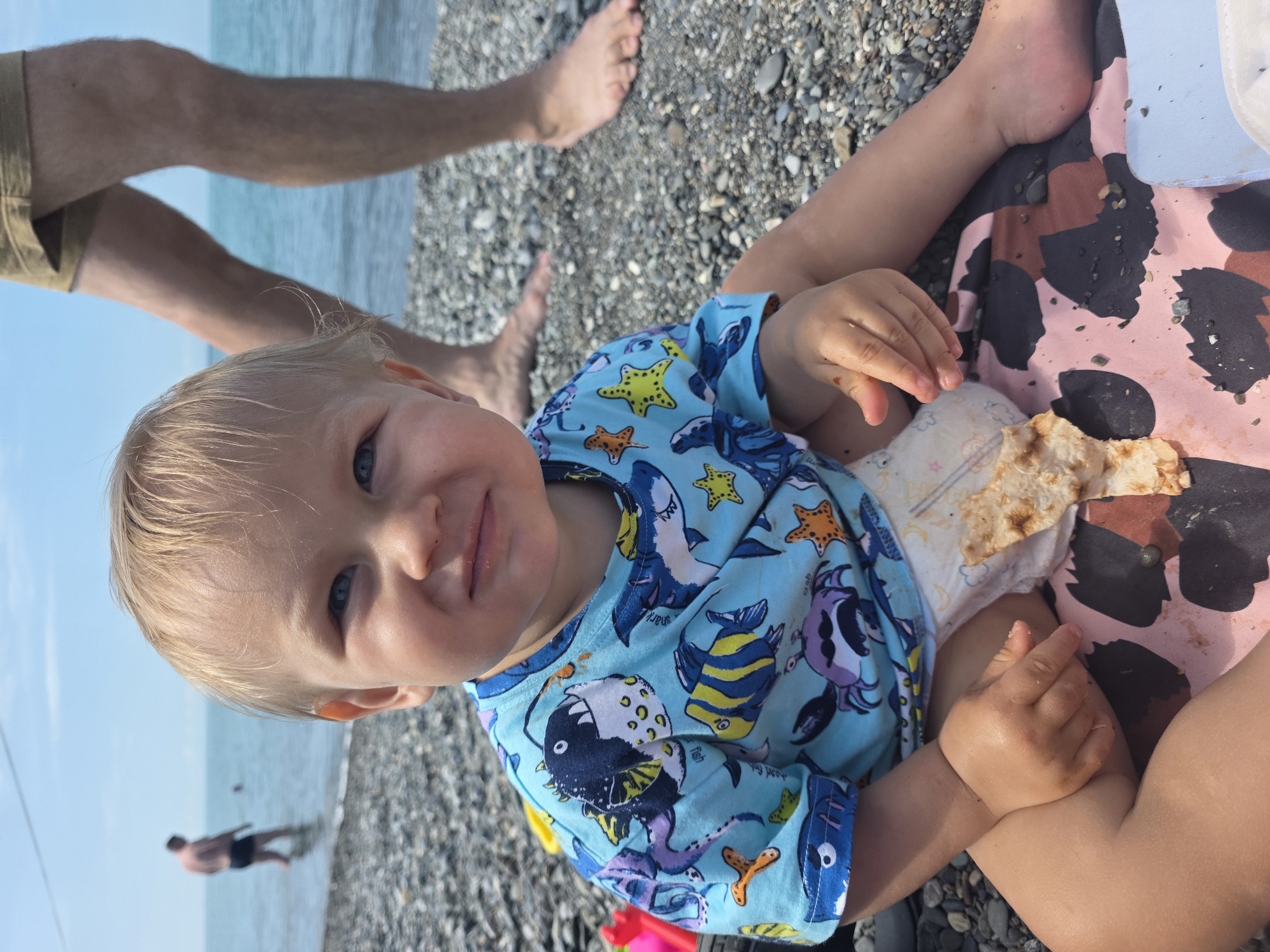 A toddler sits on a pebble beach, wearing a blue t-shirt with a fish pattern and a diaper. A piece of flatbread rests on the diaper. The toddler is facing the camera, with a person's legs visible in the background. The sea is visible behind them. The scene appears to be a beach in Sochi, Russia, indicated by the presence of other people in the water and the characteristic pebble beach.