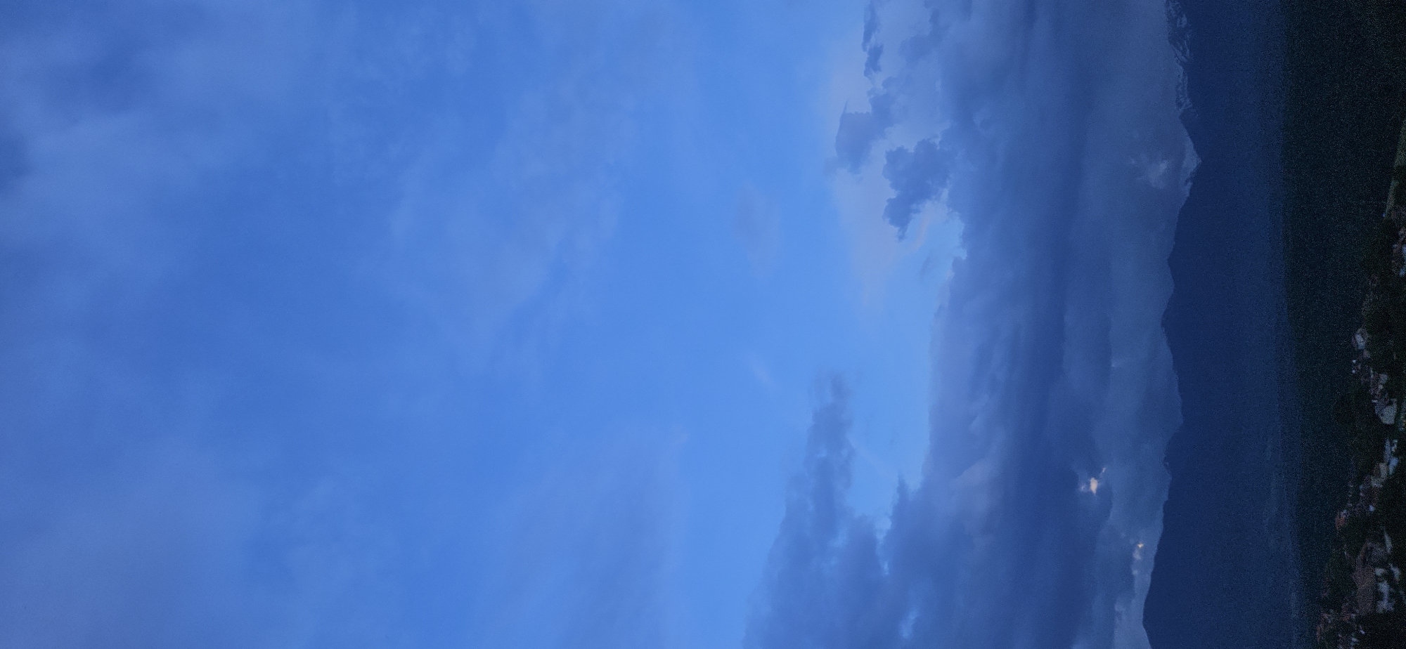 The image displays a vast natural landscape, primarily composed of a dramatic sky and a distant mountain range. The scene is oriented in a landscape format, with the sky dominating the left and central portions, and mountains appearing on the right.
The sky is filled with varying shades of blue and gray, indicating overcast conditions or the approach of dusk. On the far left, the sky is a lighter, clear blue, transitioning into areas with soft, wispy clouds. Towards the right, the clouds become denser and darker, appearing as heavy formations in deep blue and gray tones, suggesting a moody atmosphere or potential rain.
On the right side, a dark silhouette of a mountain range or large hills stretches vertically, blending into the darker clouds. The mountains are indistinct but clearly define the horizon line, their peaks appearing rugged. At the very bottom right corner, subtle hints of artificial lights or small structures are visible, suggesting a small settlement or scattered buildings nestled at the base of the mountains.
The lighting implies late afternoon, dusk, or early evening. No direct sunlight is visible, contributing to the subdued and contemplative mood of the image. The location is Gutsal, Bulgaria, placing the mountainous landscape within the context of the country's terrain. There are no identifiable people or specific objects, and no text is visible within the frame. The overall impression is one of a tranquil yet atmospheric mountainous vista under a cloudy, twilight sky.