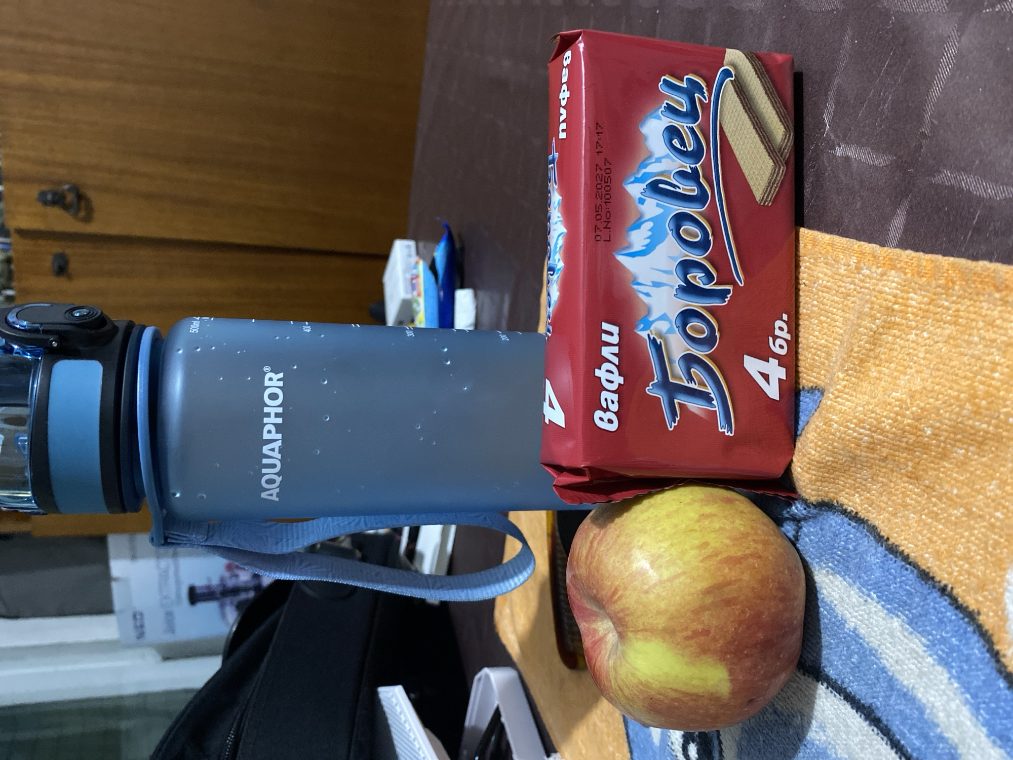 A close-up, eye-level shot shows a spread of food and a water bottle on a table. In the foreground, a ripe red and yellow apple rests on a blue and orange striped towel. To its right, a red package of wafers, branded "Боровец", is prominent, featuring an image of snow-capped mountains and the number "4" with "бр." below it. The package also displays a date and batch number: "07.05.2027 17:17 L.No:100507". Behind the apple and wafers, a blue translucent water bottle with a black and blue lid stands upright. Text on the bottle reads "AQUAPHOR" and has volume markings "500ml", "400", and "300". A blue strap is attached to the bottle's lid. In the blurred background, a dark bag is visible on the left, and wooden cabinet doors are on the right. The setting appears to be indoors, possibly a home, given the wooden furniture and the casual arrangement of objects. The image does not provide any specific visual clues to indicate the location as Smolyan, Bulgaria.