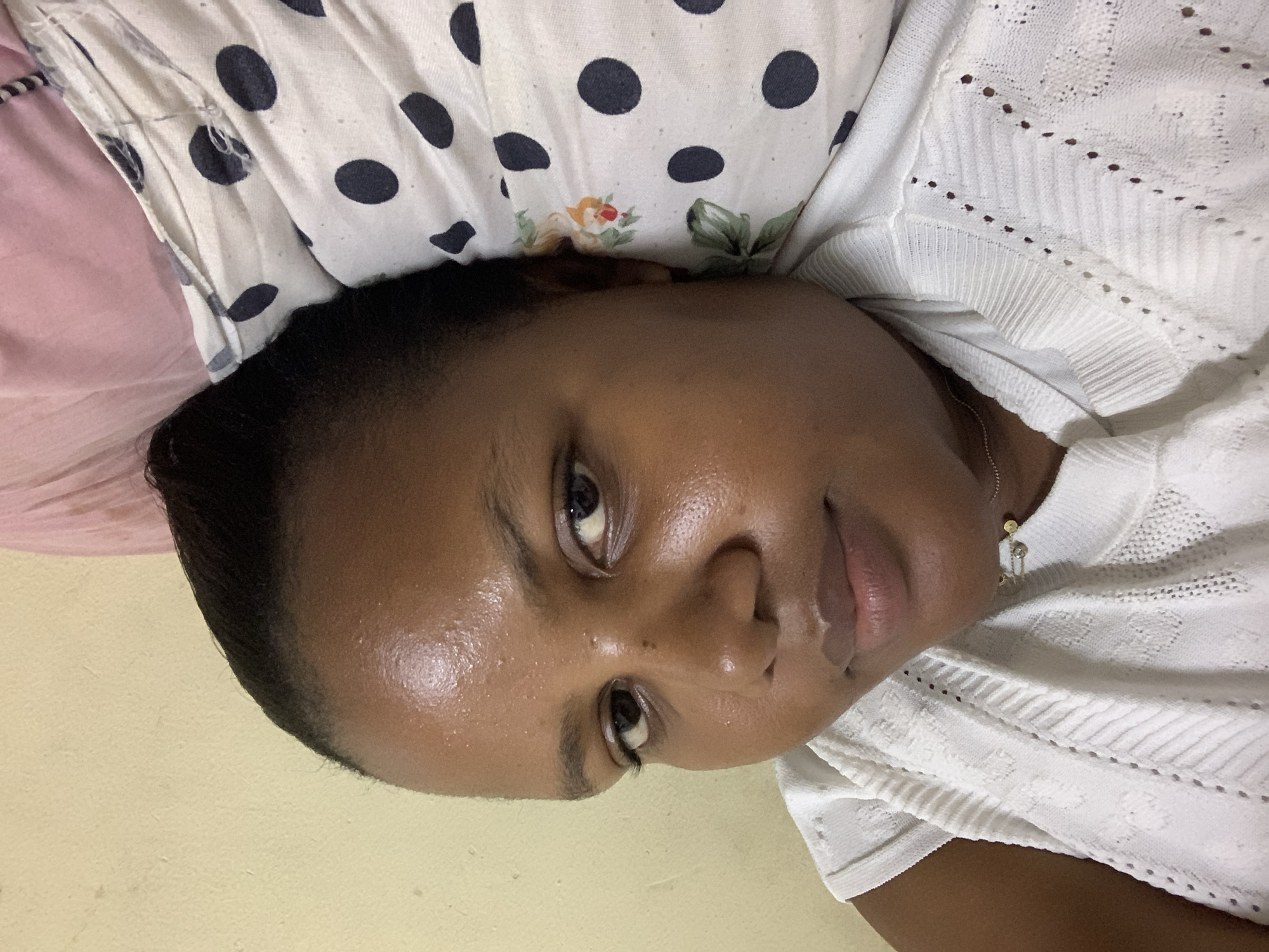 A close-up, portrait-style image shows a woman with dark hair and skin lying on her side. Her head is slightly tilted toward the camera, and she has a neutral expression. She is wearing a white, textured top and a delicate necklace with small charms. Beneath her head is a pillow or cushion with a white background and black polka dots, with floral accents visible as well. The background appears to be a plain, light-colored wall. The image does not provide any information to confirm that it was taken in Dar es-Salaam, Tanzania.
