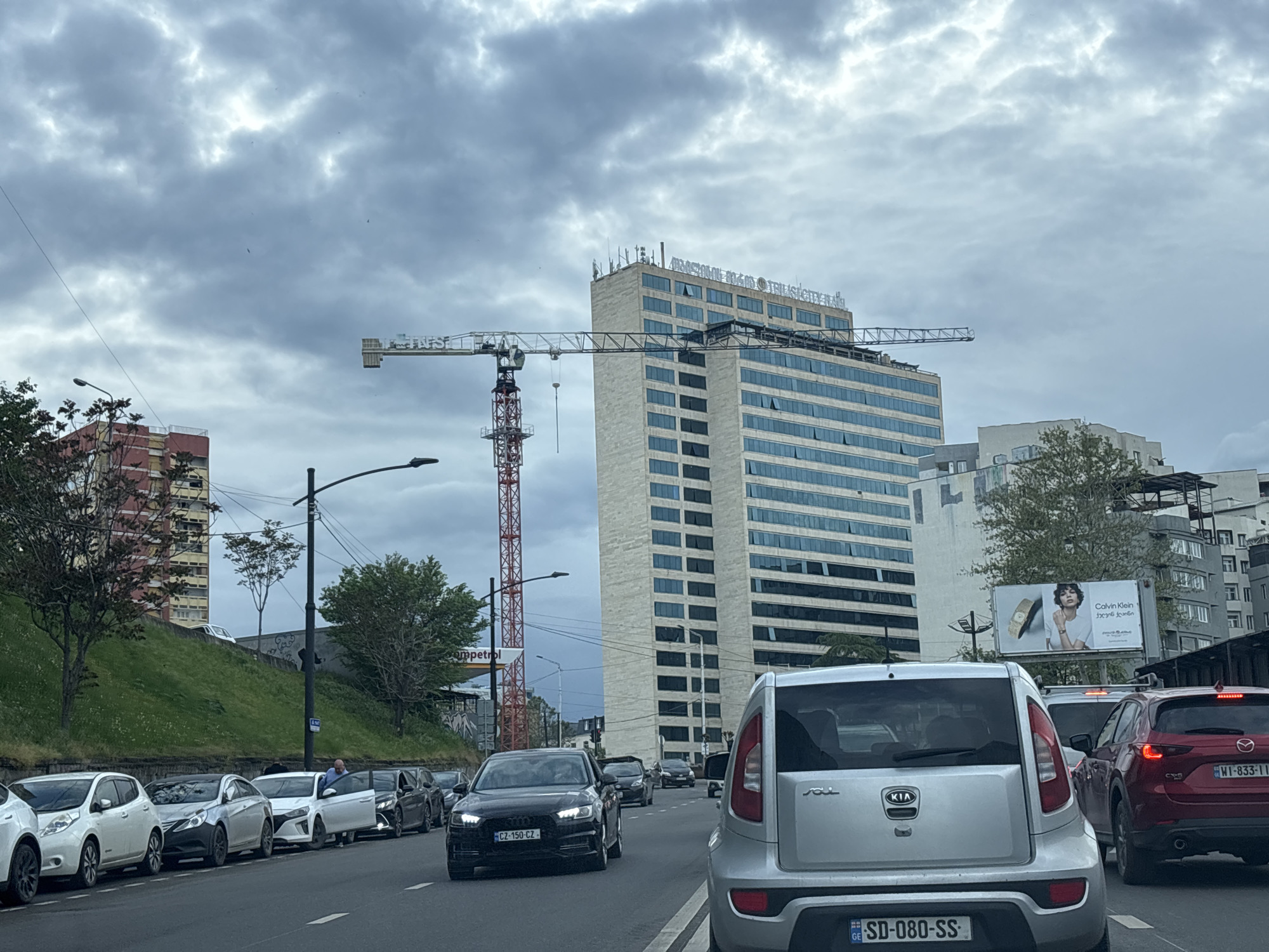 The image captures a street scene in Tbilisi, Georgia, characterized by heavy traffic on a cloudy day. Dominating the background is the Axis Towers complex, with a construction crane visible nearby. A billboard promoting Calvin Klein is to the right of the tower. Several cars are visible, with license plates from Georgia, the Czech Republic, and Poland. In the foreground, cars are densely packed on the road. The license plate of the silver Kia Soul reads "SD-080-SS." A red Mazda CX-5 is also visible on the right with the license plate "WI-833-11." A black Audi displays the license plate "CZ-150-CZ-." A man is partially visible in a parked car on the left side of the road. A gas station sign is partially visible in the background.