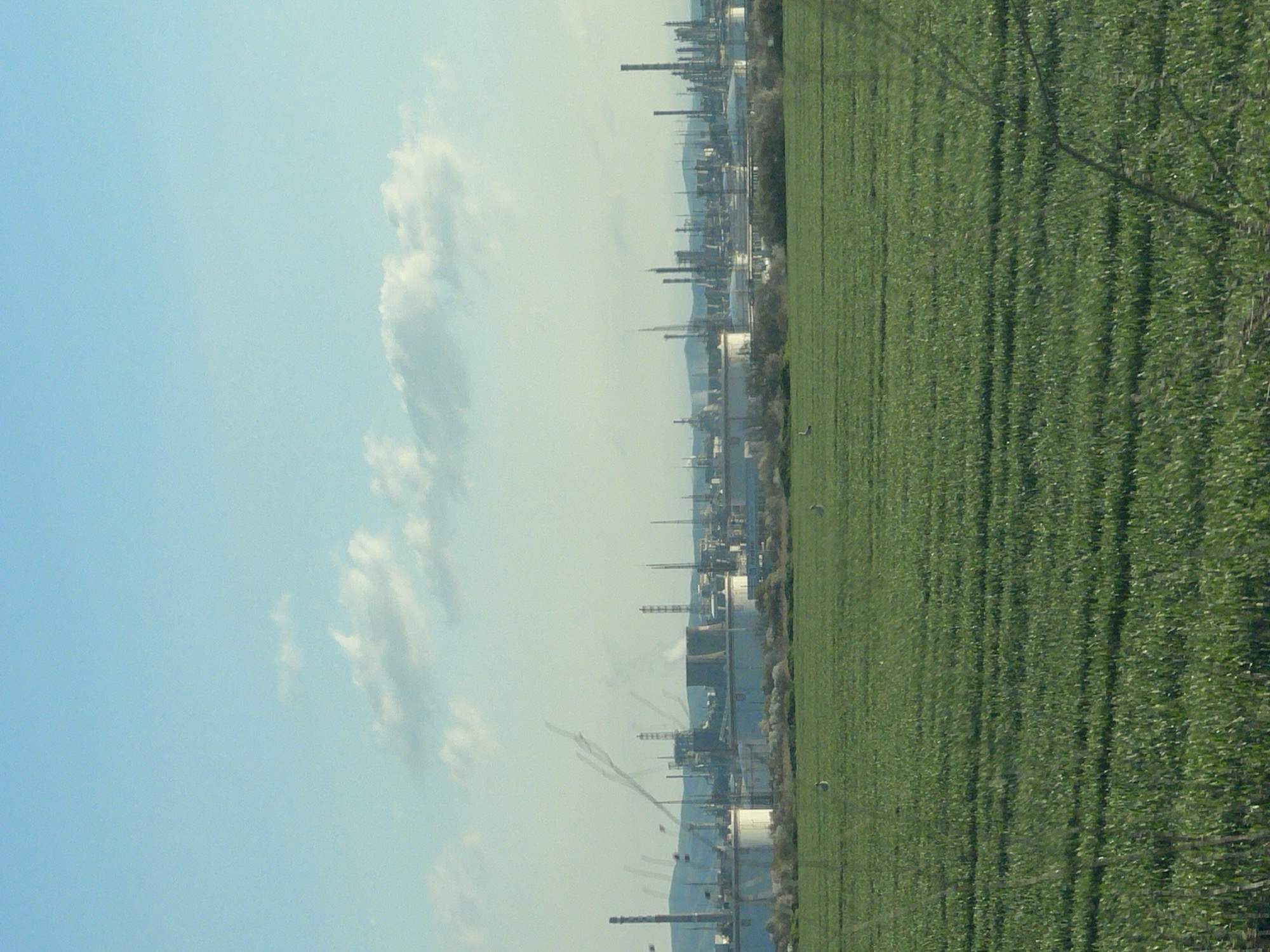 The image shows a landscape with a green field on the right and a clear blue sky with some white clouds on the left. In the middle, there is a skyline of industrial structures, possibly factories or refineries, suggesting the presence of an industrial zone. The location is Bratovo, Bulgaria.