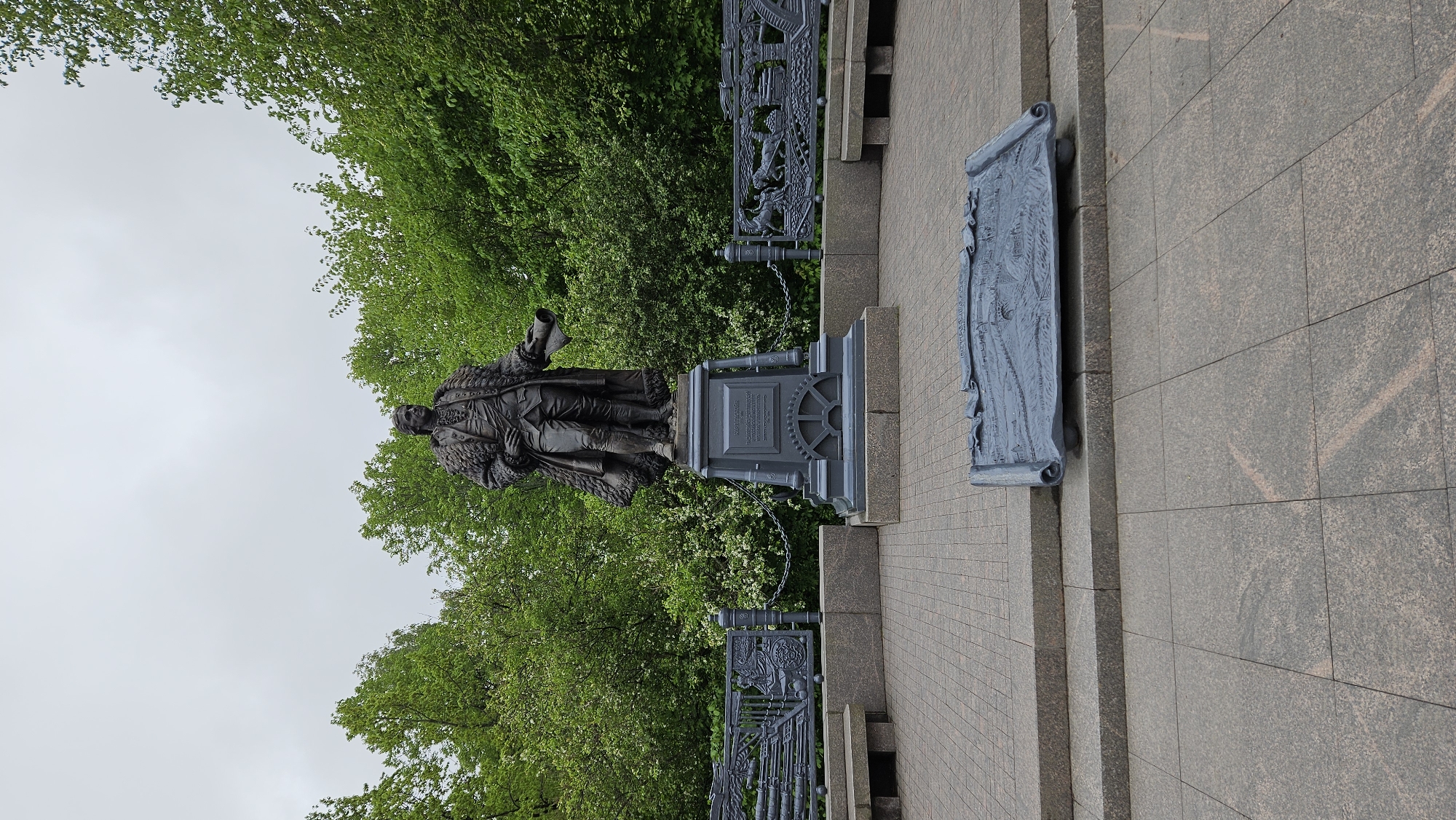 The image shows the Alexander von Humboldt monument in Berlin, Germany. The statue of Humboldt stands on a stone pedestal surrounded by greenery and a decorative iron fence. A sculpted scroll or plaque is mounted to the side of the monument. The sky is overcast.
