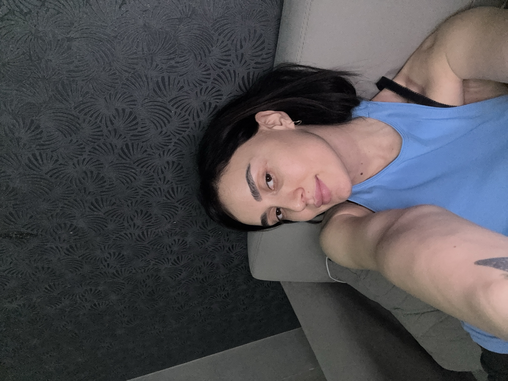 A medium shot, taken from above and to the side, shows a person reclining on a gray sofa in front of a patterned dark gray wall. The person is wearing a blue tank top and has dark hair and fair skin. Their face is slightly tilted towards the camera. A black bra strap is visible under the tank top. Part of an arm with a tattoo is visible in the foreground. The location cannot be determined from the given information.